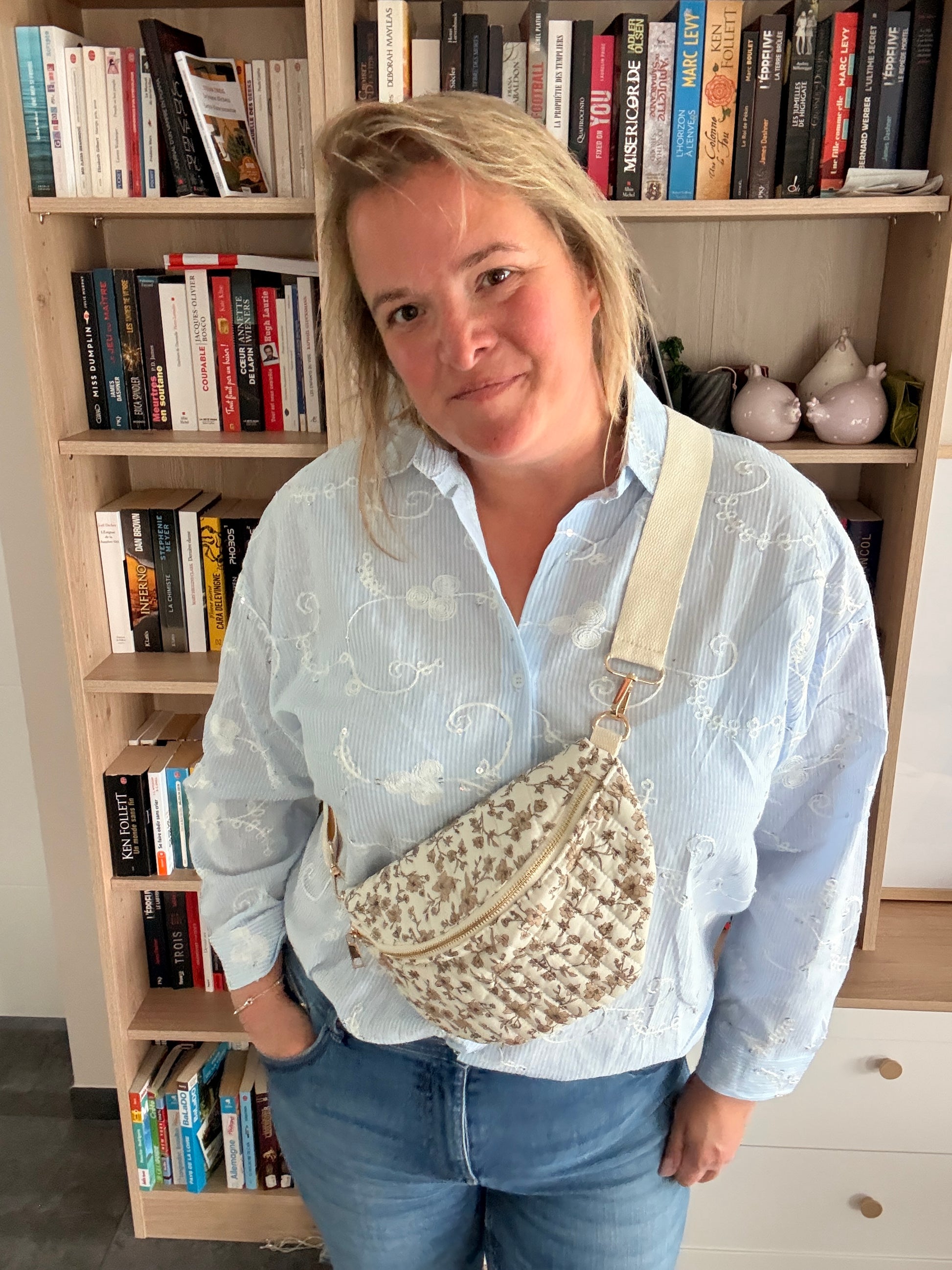 Sac banane femme fleurie taupe avec fond ivoire porté en bandoulière réglable, doré zip et look casual devant étagère de livres.