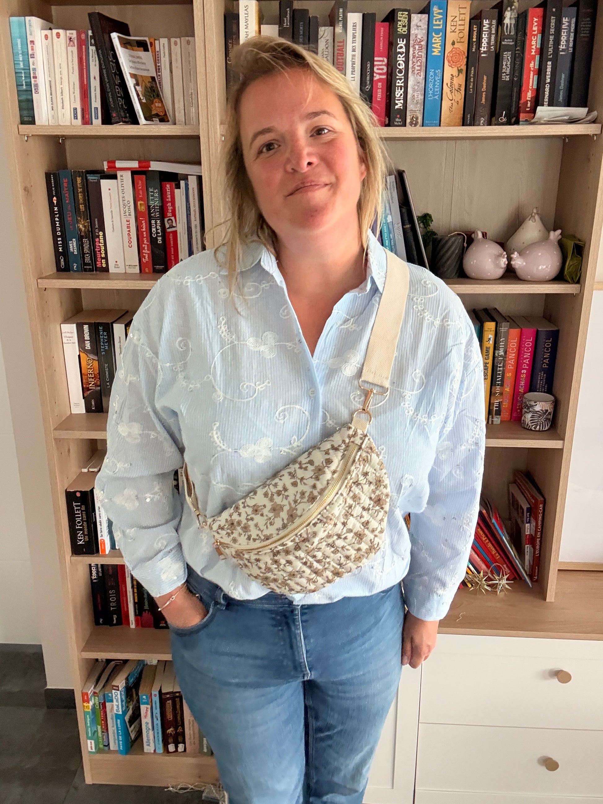 Sac banane femme fleuri taupe fond ivoire avec zip doré porté en bandoulière réglable sur une femme devant une bibliothèque.