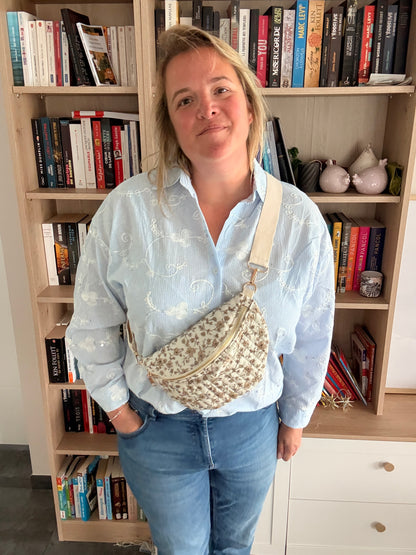 Sac banane femme fleuri taupe fond ivoire avec zip doré porté en bandoulière réglable sur une femme devant une bibliothèque.