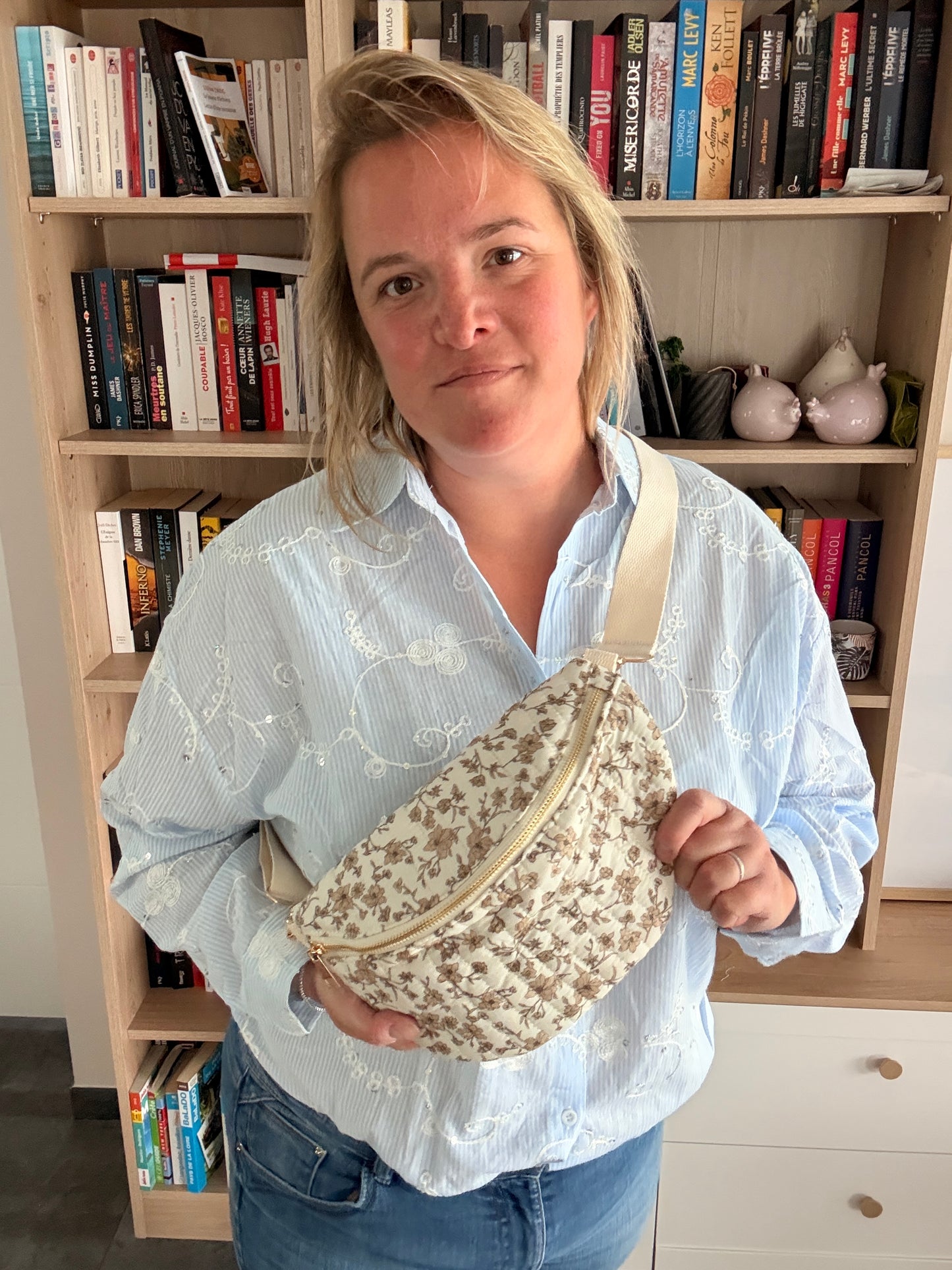 Femme portant un sac banane fleuri taupe avec fond ivoire, zippé doré et bandoulière ajustable, debout devant bibliothèque.