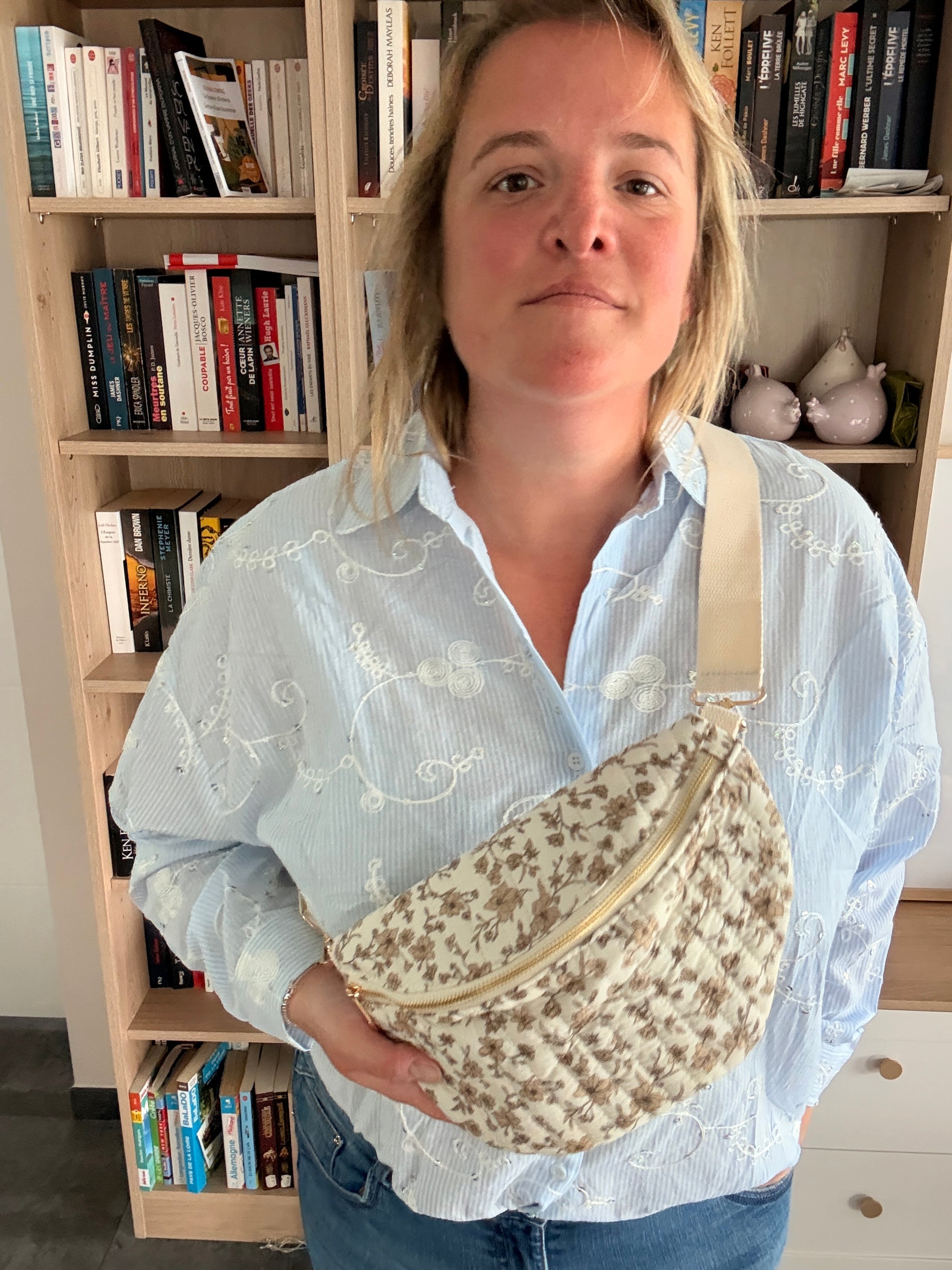 Sac banane femme fleuri taupe fond ivoire, zip doré et bandoulière réglable portée sur l’épaule, femme en chemise bleu clair devant une étagère de livres.