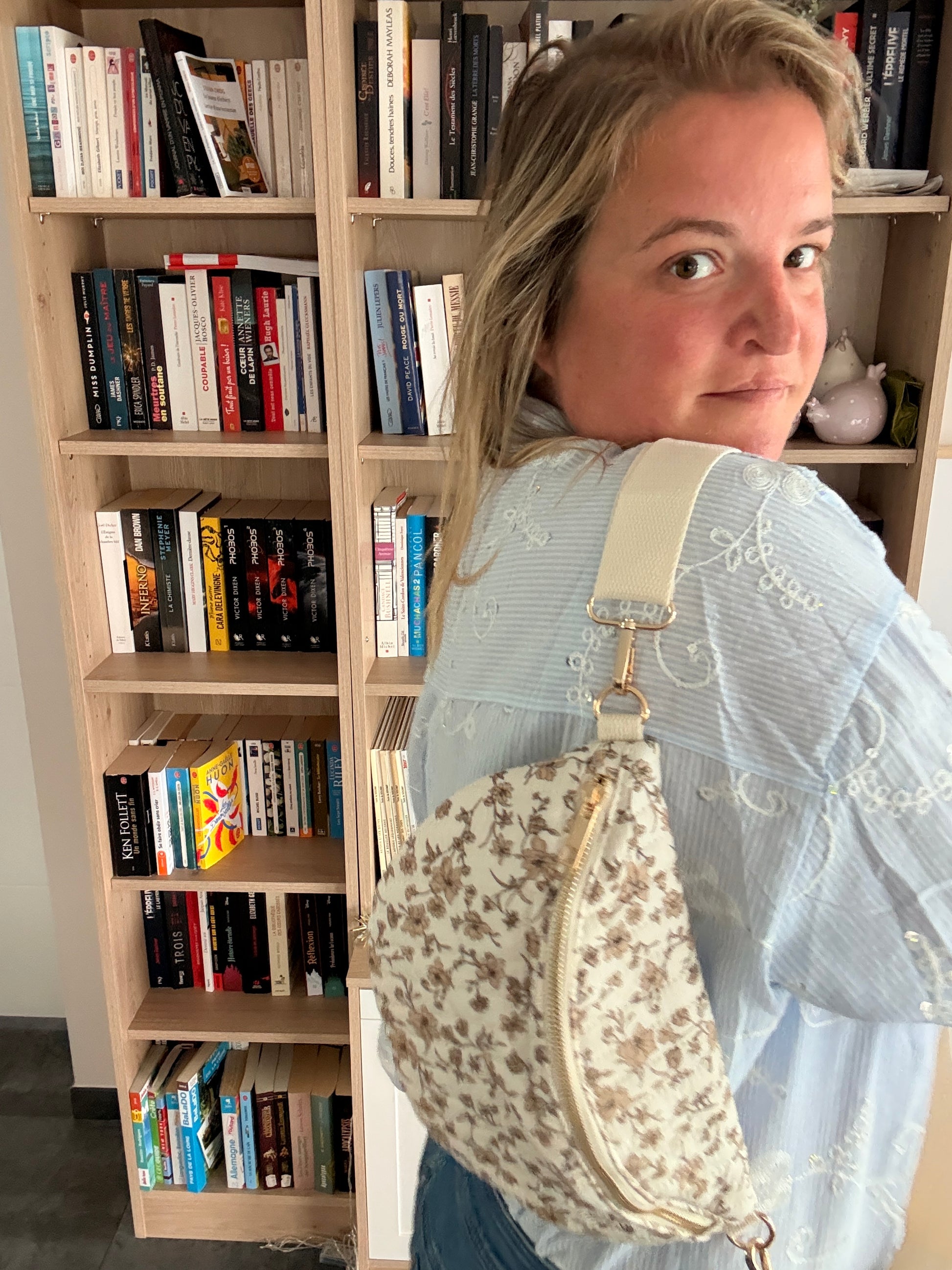 Sac banane femme fleurie taupe à fond ivoire porté en bandoulière réglable, avec zip doré, devant une bibliothèque.