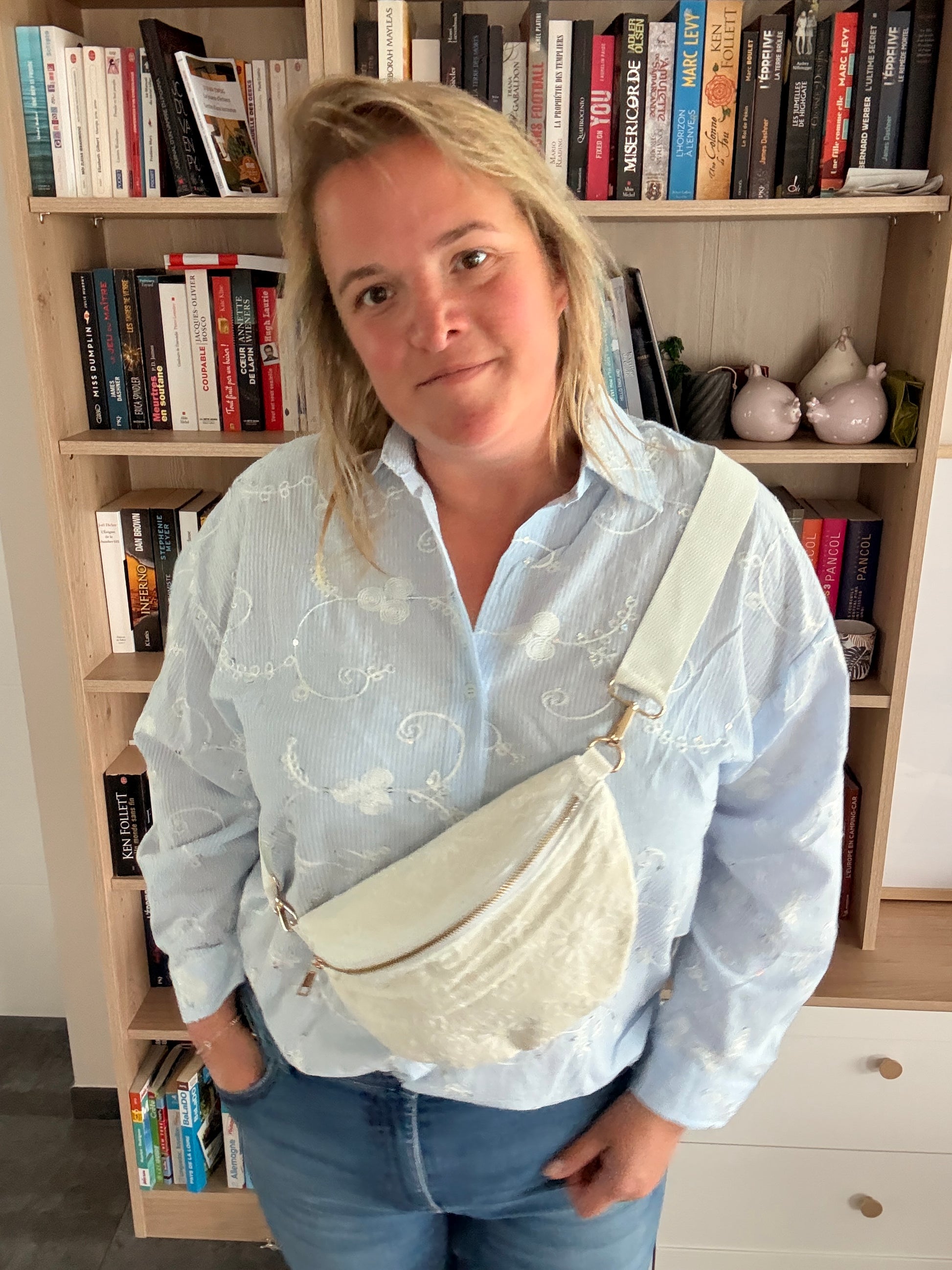 Sac banane blanc floral irisé porté en bandoulière sur l’épaule droite, portée par une femme en chemise bleue, debout devant une étagère de livres.