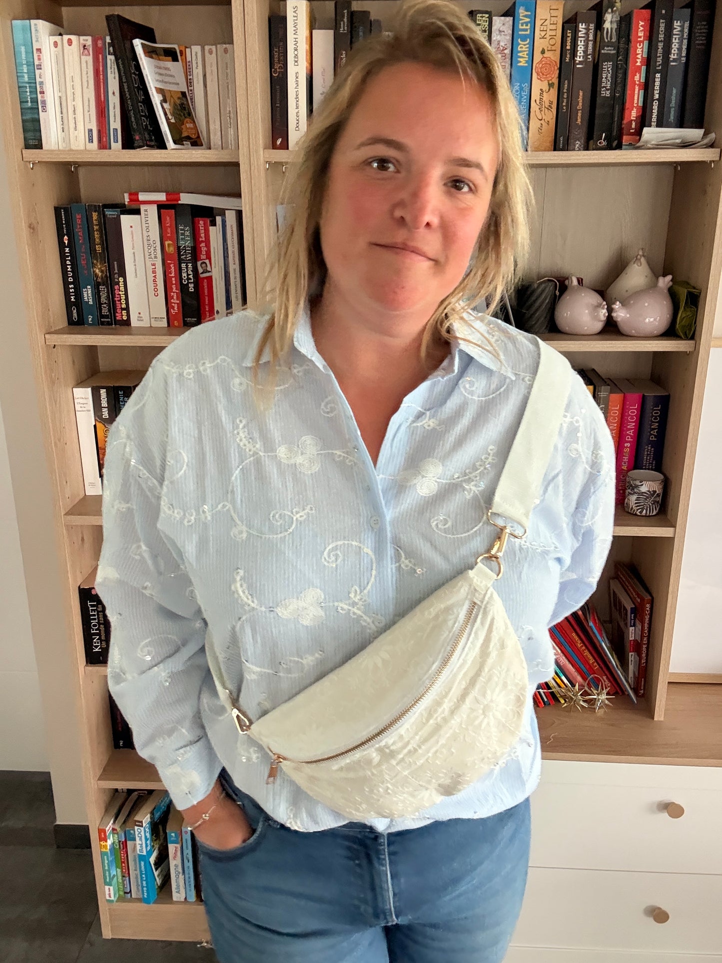 Homme ou femme portant une sac banane blanc floral irisé portée en bandoulière large et zip doré, debout devant une bibliothèque.