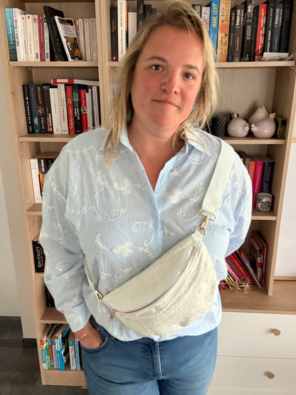 Homme ou femme portant une sac banane blanc floral irisé portée en bandoulière large et zip doré, debout devant une bibliothèque.