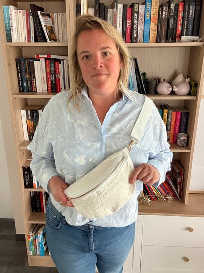 Sac banane féminin blanc avec motif florale irisée porté en bandoulière large et fermeture dorée, sur fond bibliothèque.
