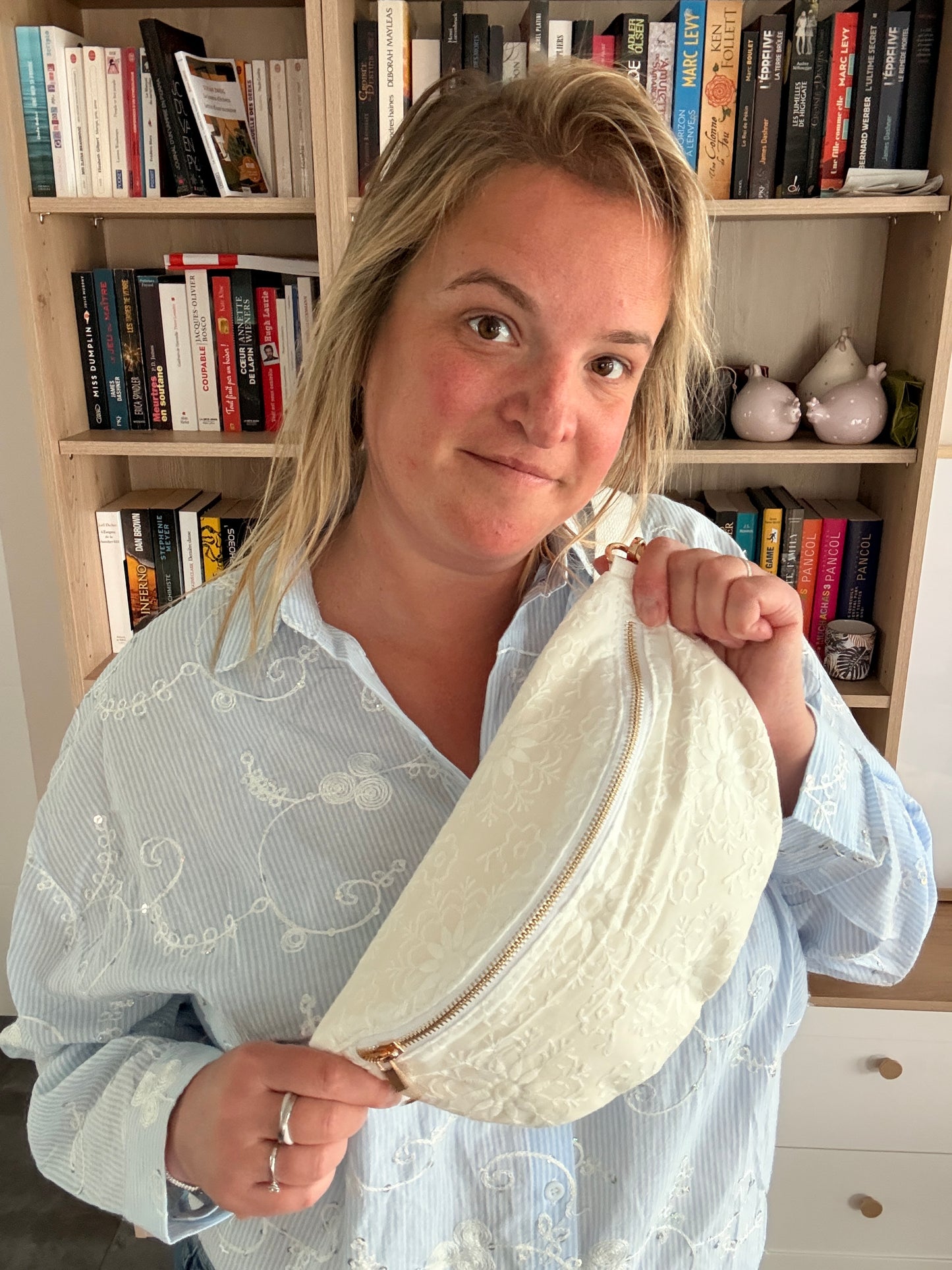 Femme tenant un sac banane blanc floral irisé avec bandoulière large et fermeture dorée, debout près d'une étagère de livres.