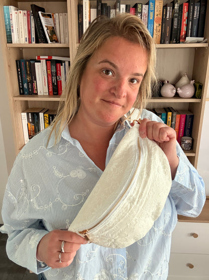 Femme tenant un sac banane blanc floral irisé avec bandoulière large et fermeture dorée, debout près d'une étagère de livres.