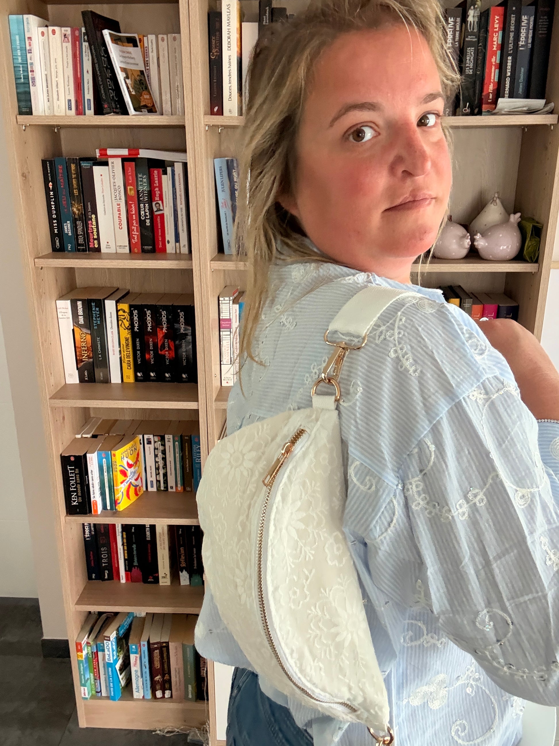 Sac banane blanc floral irisé porté en bandoulière par une femme, devant une étagère de livres.