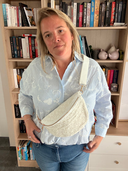 Sac banane femme en broderie anglaise crème portée en bandoulière, look romantique-bohème devant une bibliothèque.