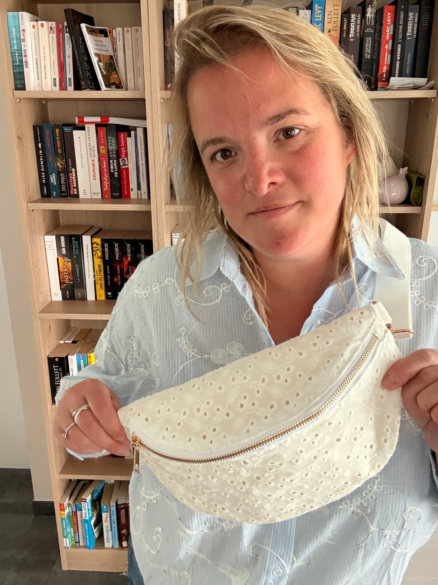 sac banane femme en broderie anglaise crème, porté par une femme devant une étagère de livres; elle tient la sangle zippée en coton brodé.