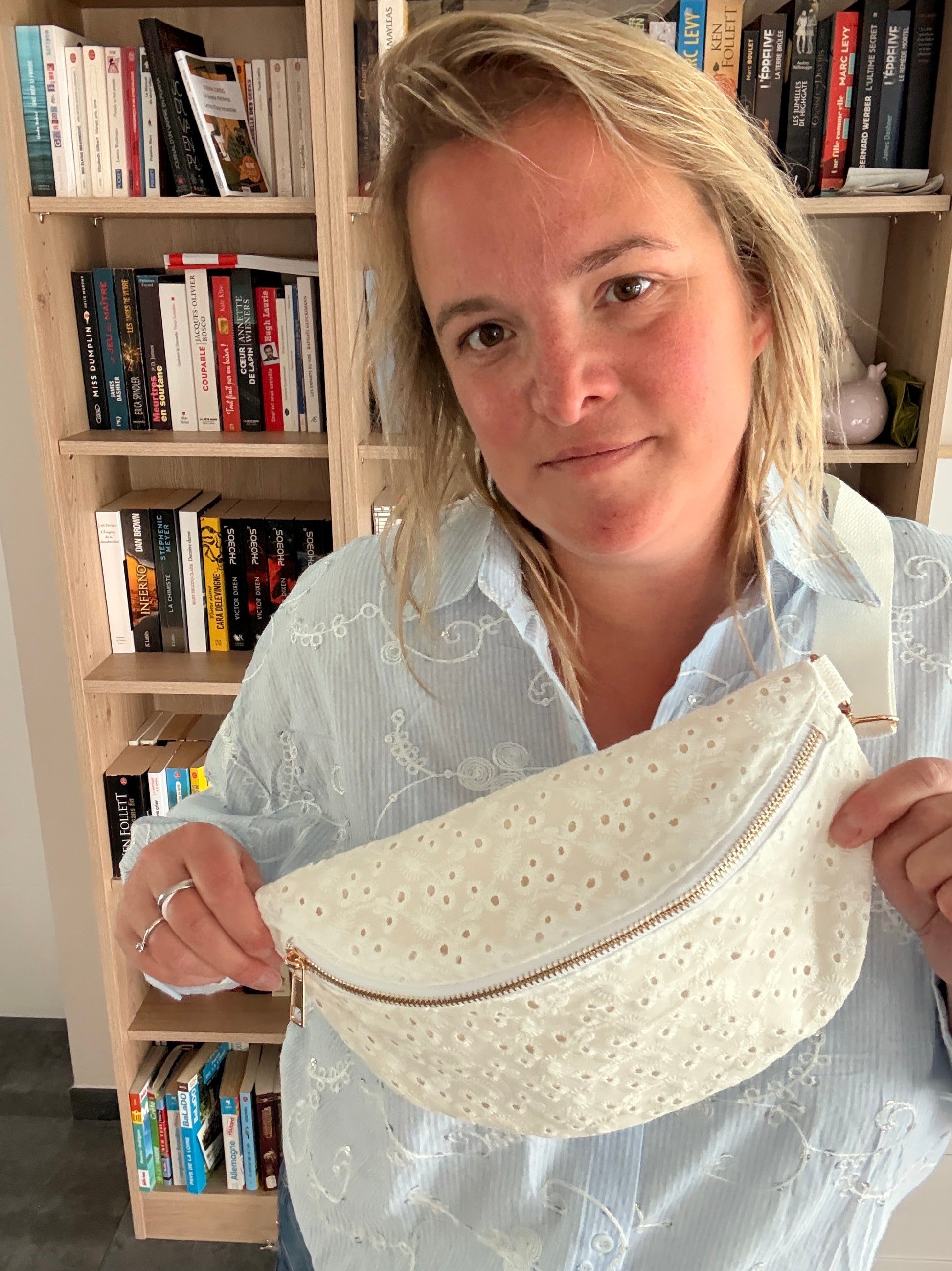 sac banane femme en broderie anglaise crème, porté par une femme devant une étagère de livres; elle tient la sangle zippée en coton brodé.