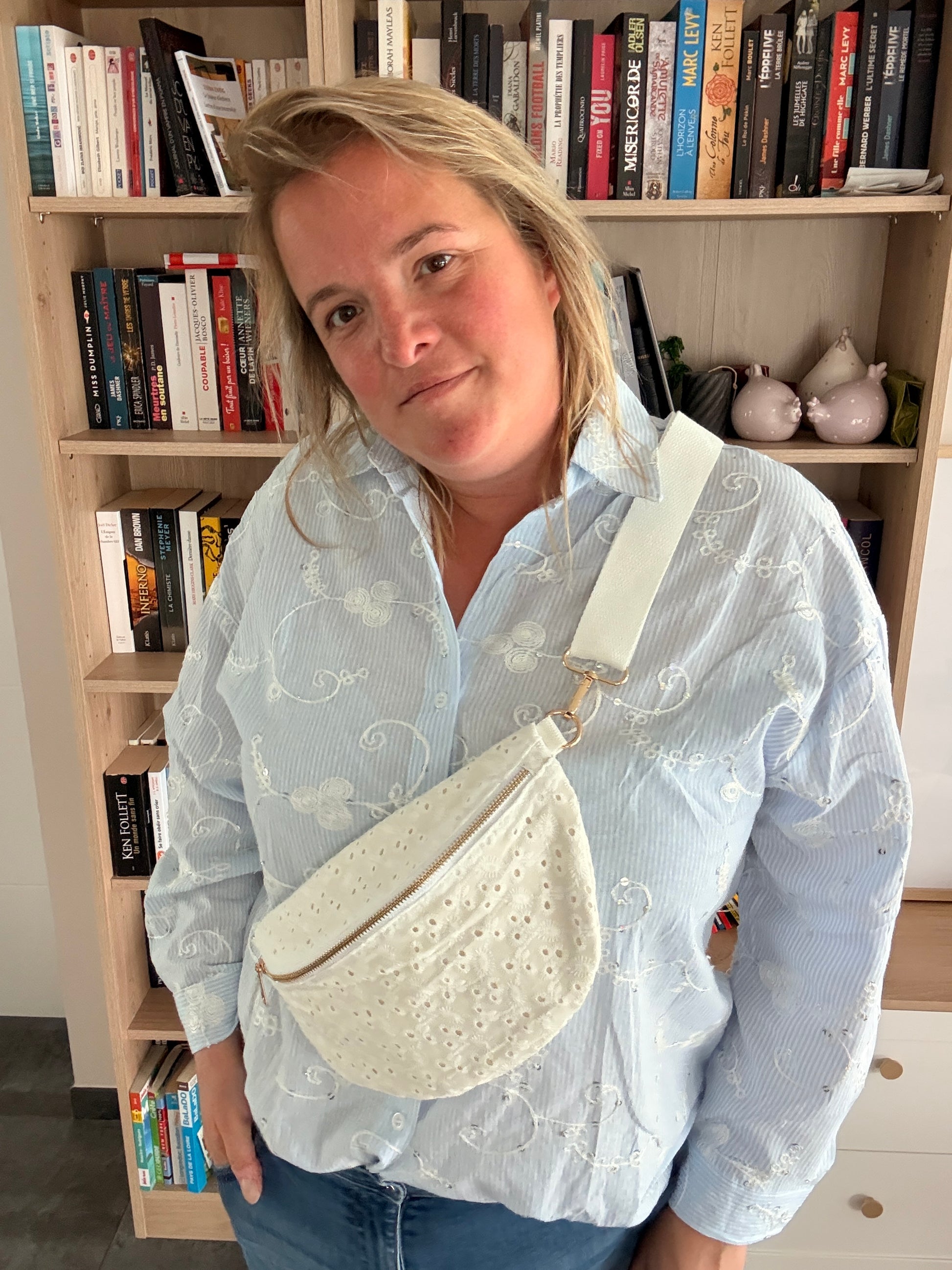 Sac banane femme en broderie anglaise crème portée sur l’épaule, sur fond bibliothèque; style romantique et bohème.