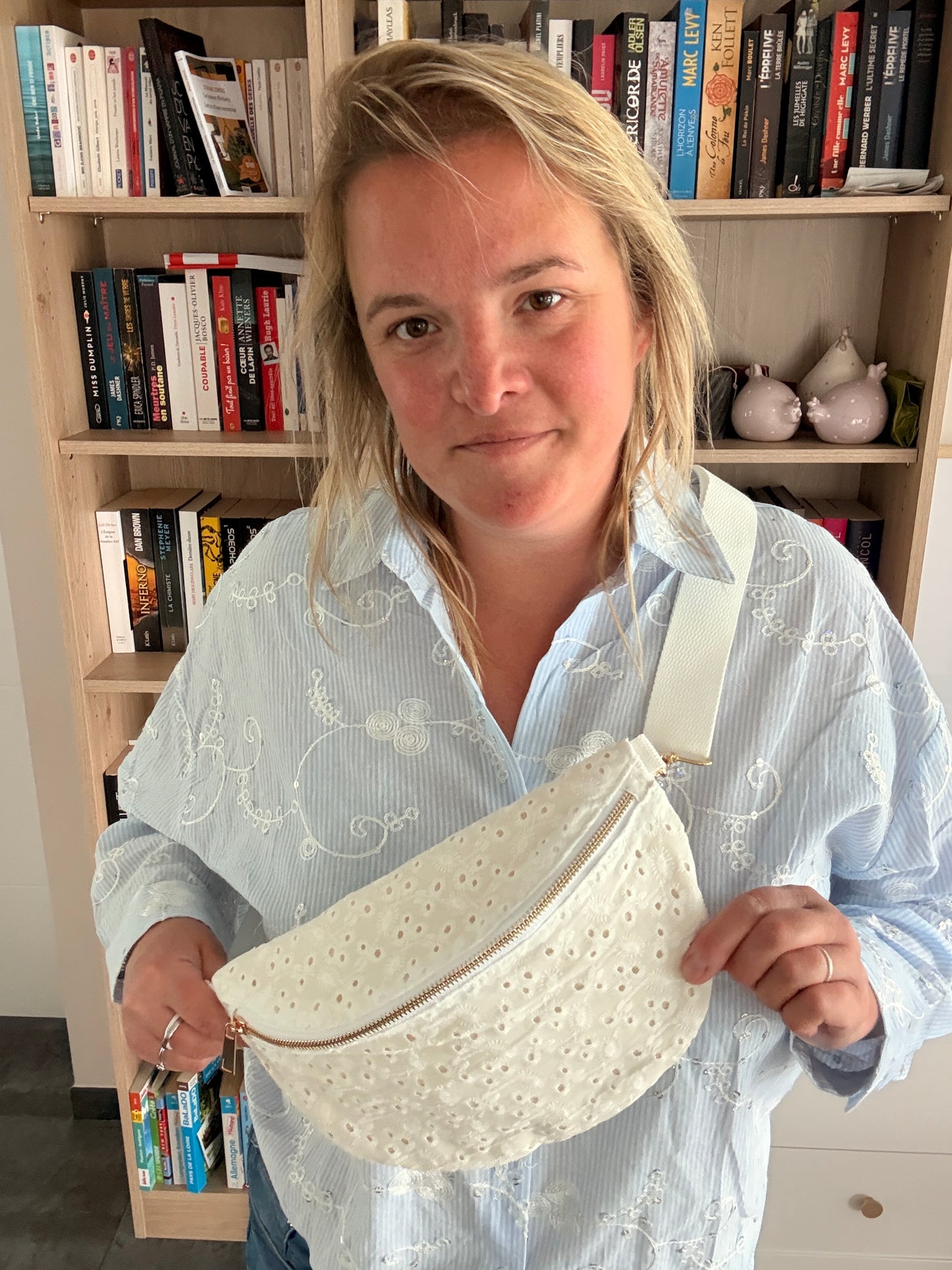 Sac banane femme en broderie anglaise crème portée en bandoulière sur une tenue décontractée, près d’une bibliothèque.