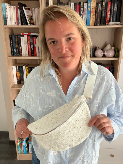 Sac banane femme en broderie anglaise crème portée en bandoulière sur une tenue décontractée, près d’une bibliothèque.
