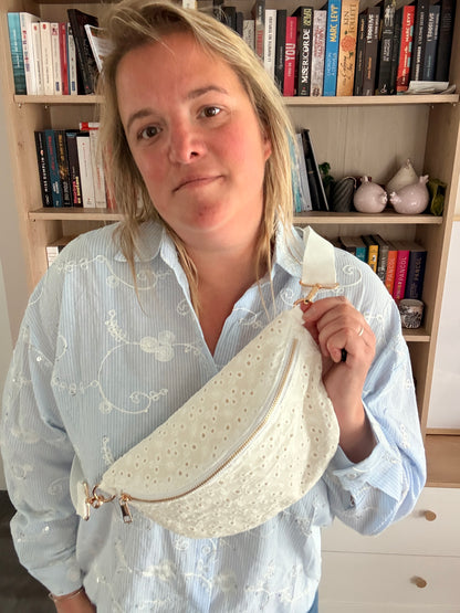 Sac banane femme en broderie anglaise crème portée en bandoulière, sur fond de bibliothèque.