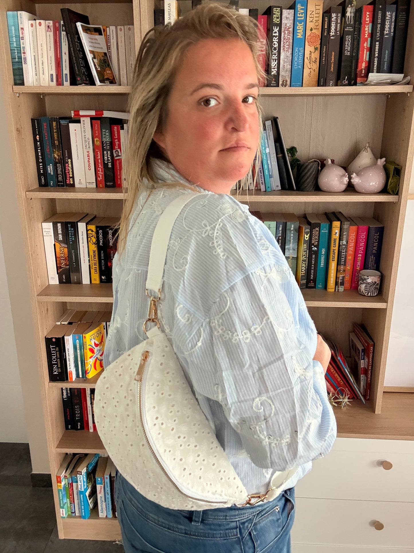 Sac banane femme en broderie anglaise crème portée en bandoulière, près des étagères et livres.
