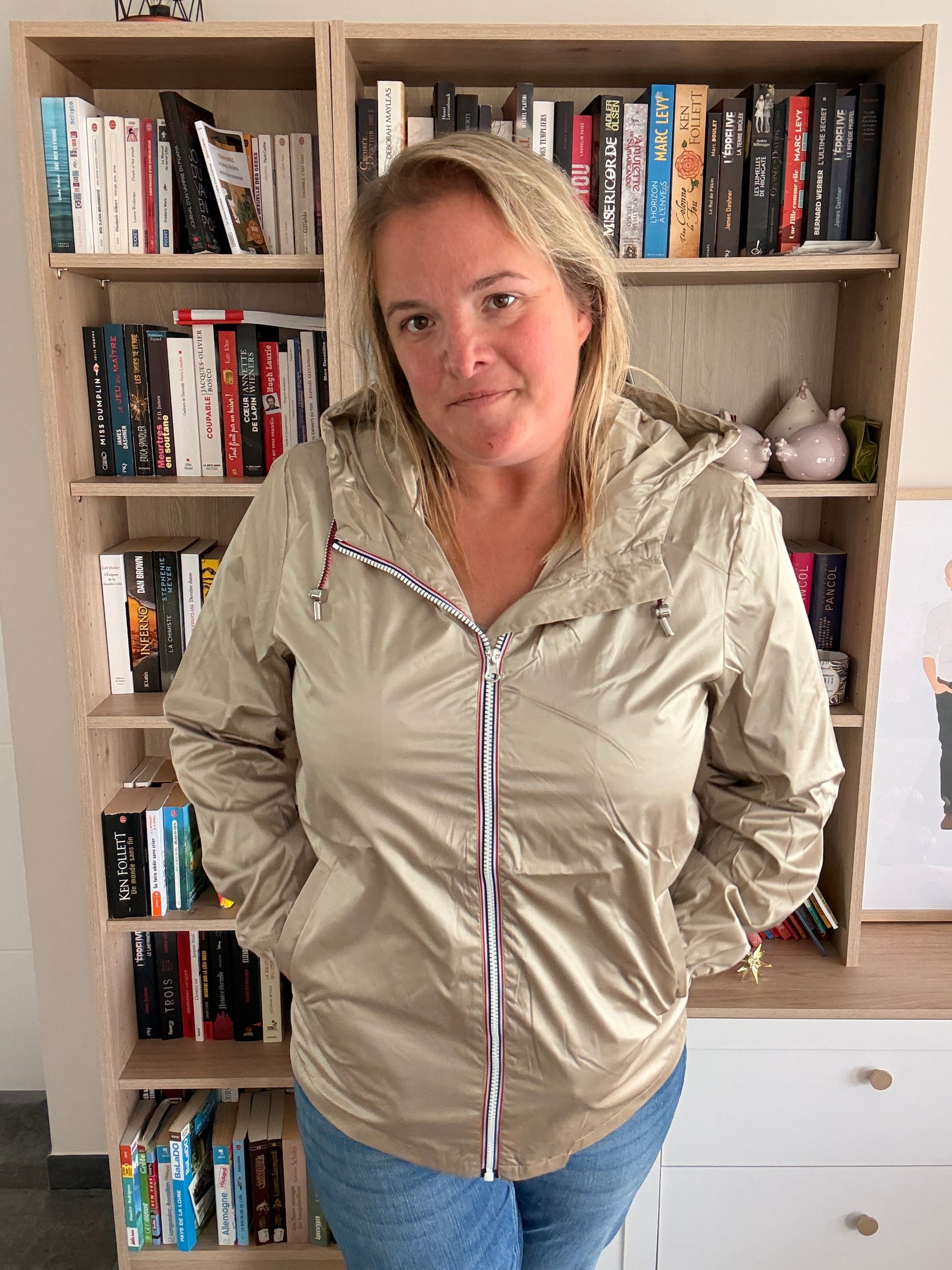 Imperméable mi-saison couleur sable irisée porté par une femme debout devant une bibliothèque.