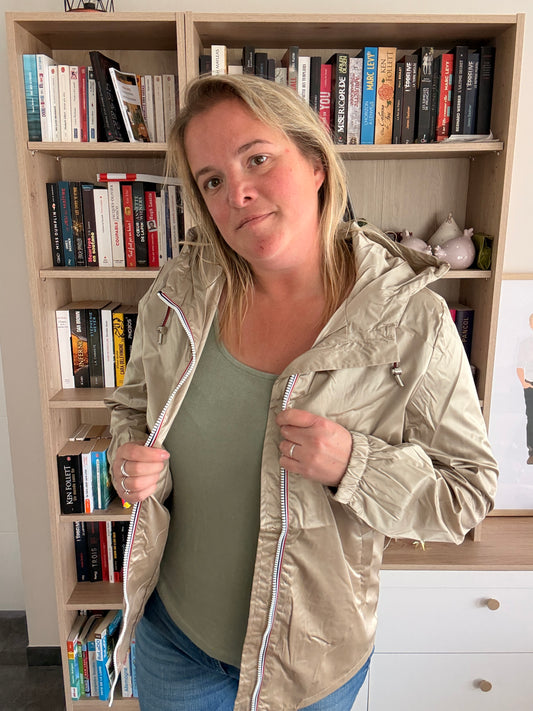 Imperméable mi-saison mi-long couleur sable irisée porté par une femme devant une bibliothèque, zippé partiellement.