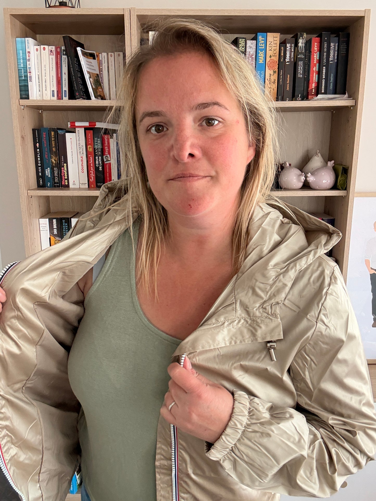 Laura Jo Imperméable Mi-Saison Mi-longue couleur Sable Irisé porté posé de côté, femme devant une bibliothèque, montrant l’imperméable beige clair.