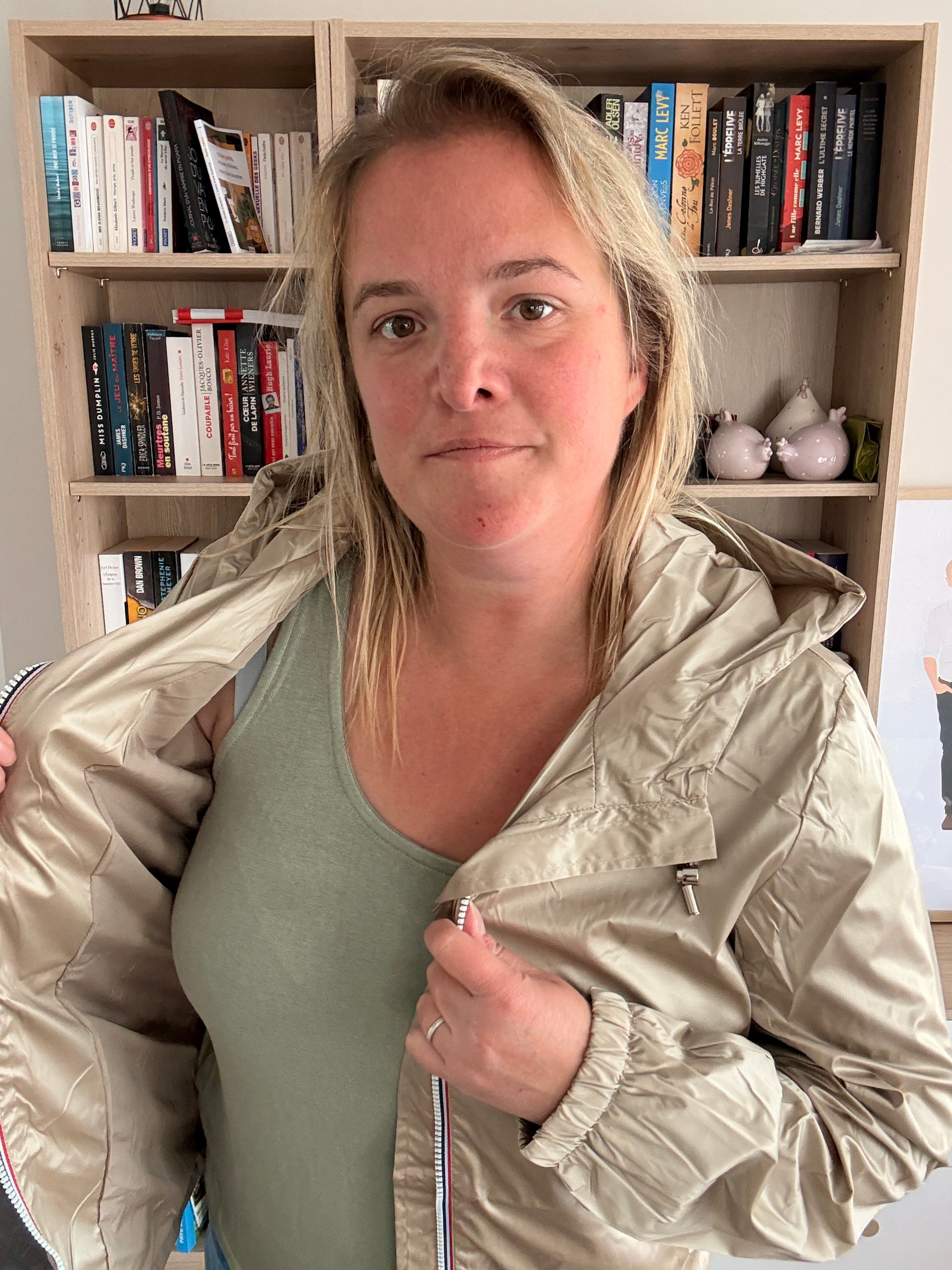 Laura Jo Imperméable Mi-Saison Mi-longue couleur Sable Irisé porté posé de côté, femme devant une bibliothèque, montrant l’imperméable beige clair.