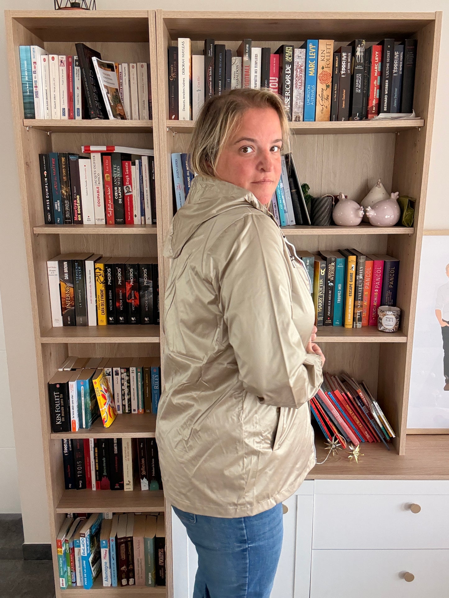 Laura Jo Imperméable Mi-Saison Mi-longue coupe-vent couleur sable irisé porté par une femme debout de profil, bibliothèque en arrière-plan.