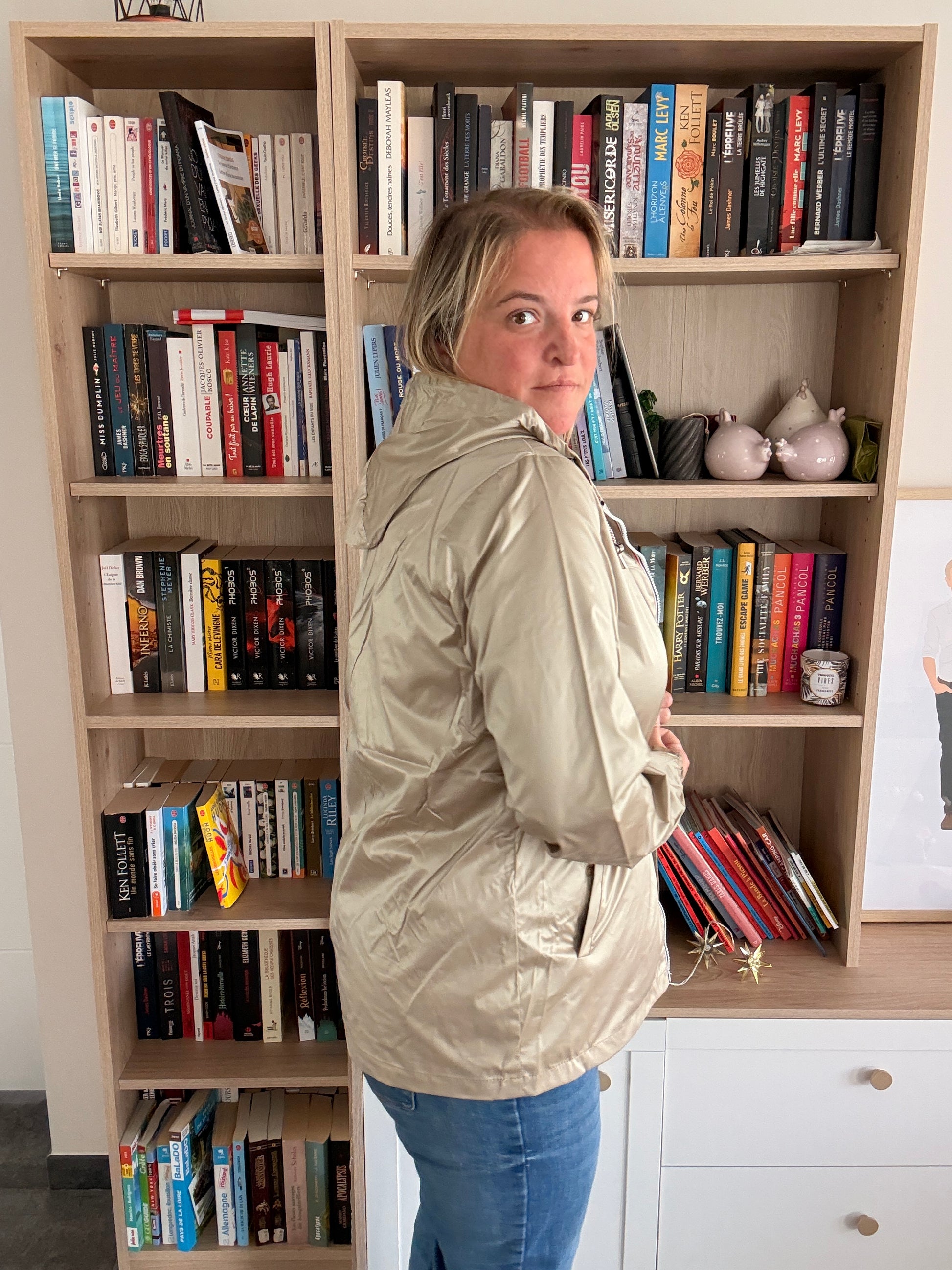 Laura Jo Imperméable Mi-Saison Mi-longue coupe-vent couleur sable irisé porté par une femme debout de profil, bibliothèque en arrière-plan.
