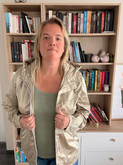 Homme ou femme portant un imperméable mi-saison couleur sable irisé, debout à l’intérieur près d’une bibliothèque.