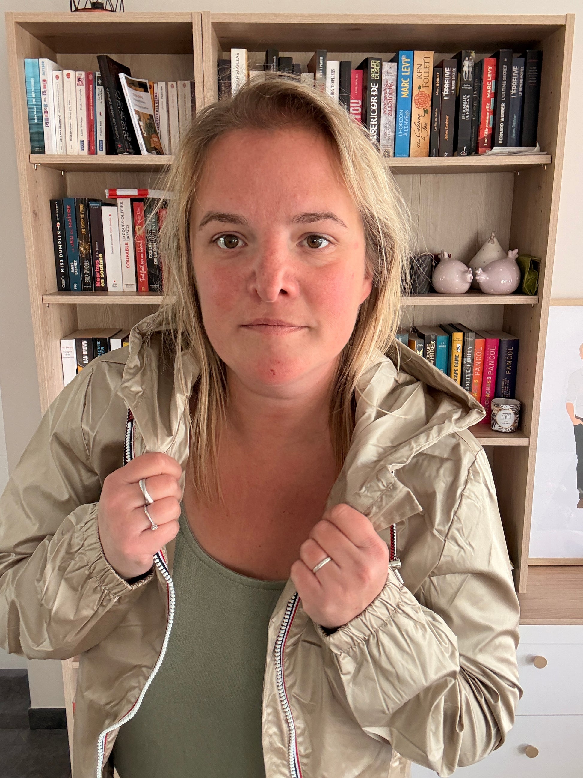 Laura Jo Imperméable Mi-Saison Mi-longue couleur sable irisé porté debout, la femme tient les bords de la veste près du col devant une bibliothèque.