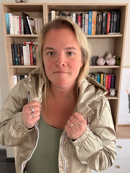 Laura Jo Imperméable Mi-Saison Mi-longue couleur sable irisé porté debout, la femme tient les bords de la veste près du col devant une bibliothèque.
