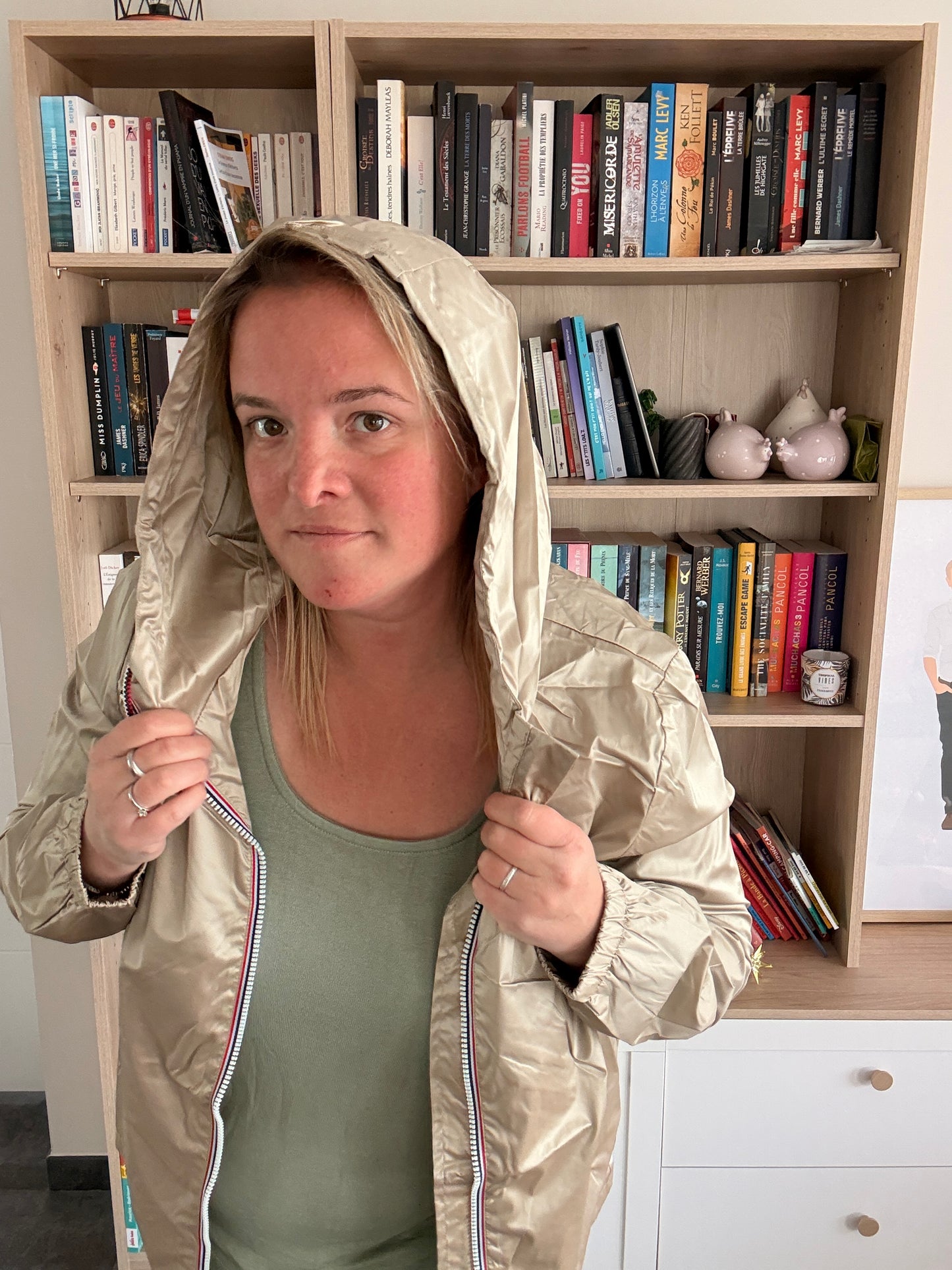 Femme portant un imperméable mi-saison couleur sable irisée, ouvrant la fermeture zippée devant elle dans une pièce avec bibliothèque en arrière-plan.