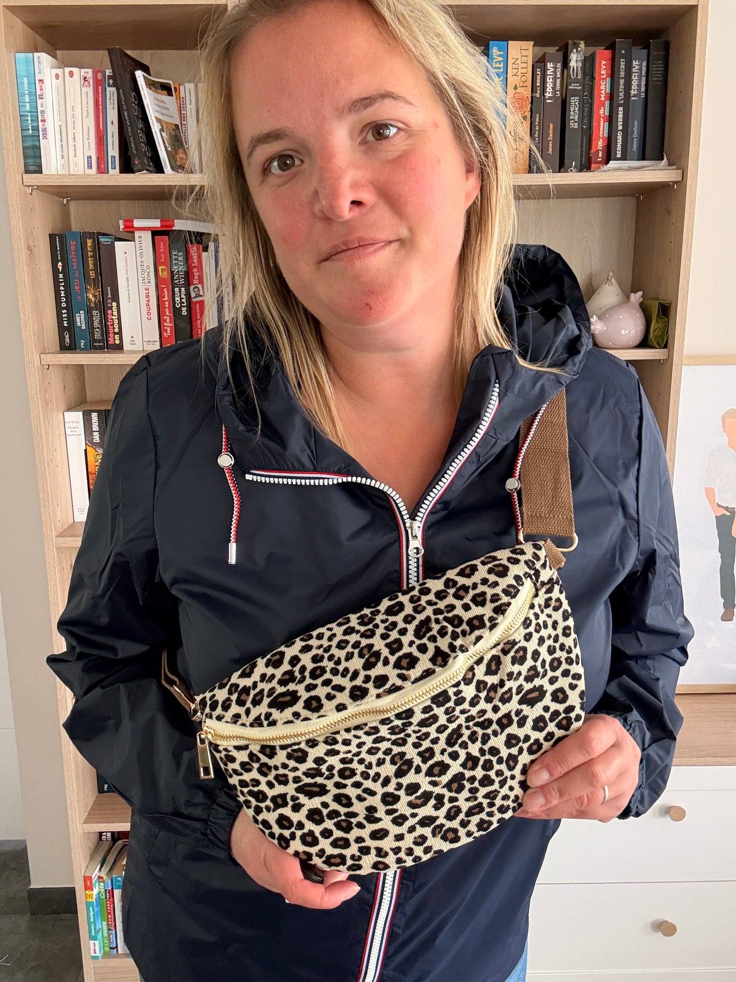 Sac banane velours côtelé imprimé léopard beige et noir porté en bandoulière à la ceinture, portée par une femme devant une bibliothèque.