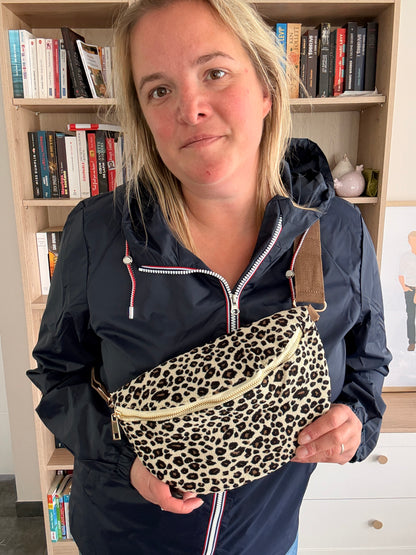 Sac banane velours côtelé imprimé léopard beige et noir porté en bandoulière à la ceinture, portée par une femme devant une bibliothèque.