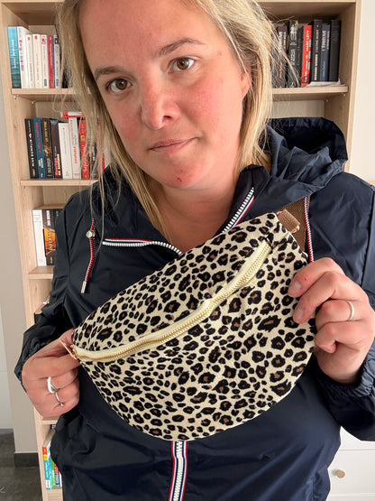 Sac banane velours côtelé imprimé léopard beige, noir et doré, porté par une femme devant bookshelf, fermeture éclair dorée.