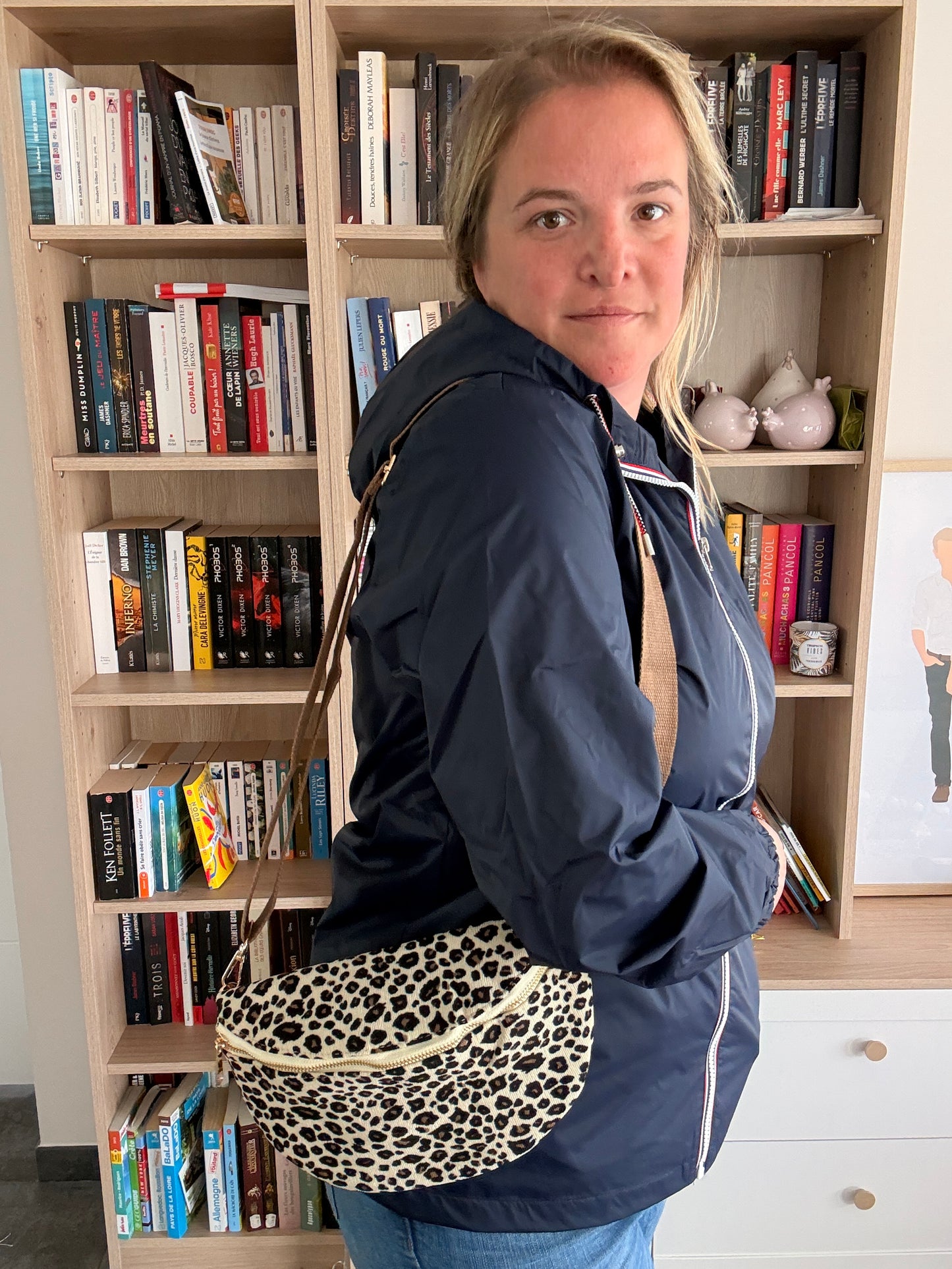 Sac banane velours côtelé imprimé léopard beige/noir/doré portée en bandoulière par une femme, près d’étagères à livres.
