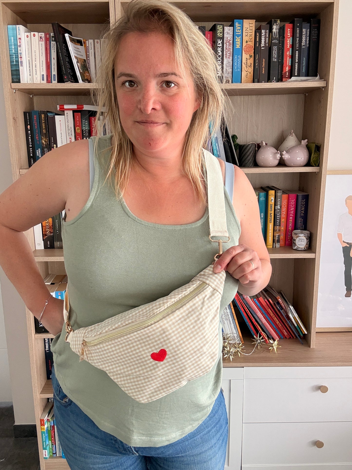 Sac banane en coton motif vichy beige et blanc avec cœur rouge porté en diagonale sur l’épaule, fond bibliothèque.