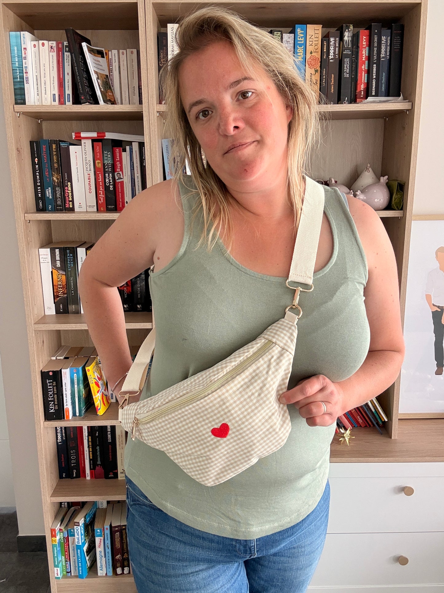 Sac banane en coton motif vichy beige et blanc avec cœur rouge porté en bandoulière par une femme dans une pièce avec bibliothèque.