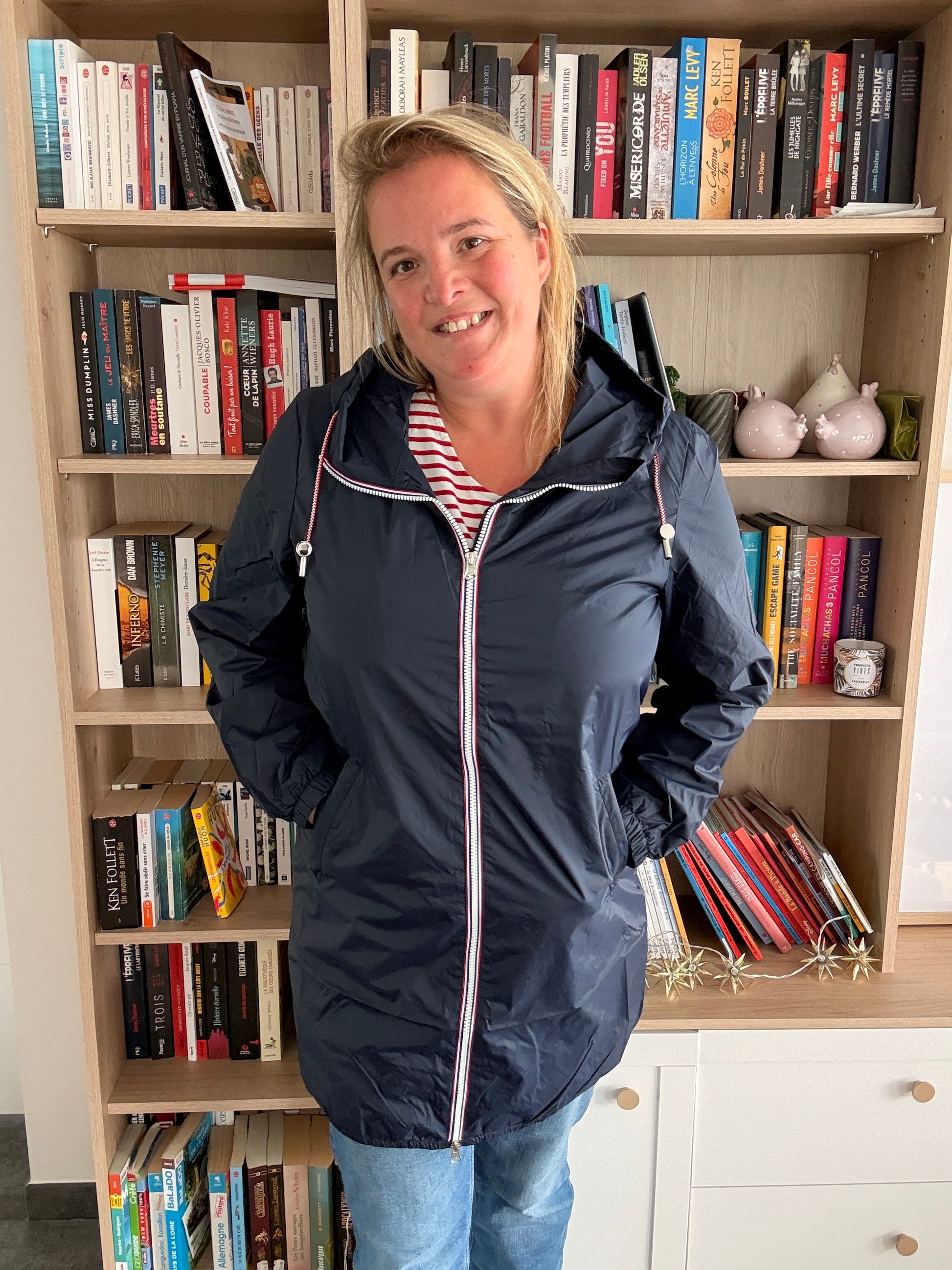 Parka manteau bleu marine imperméable long avec doublure légère, femme souriante debout devant une bibliothèque.