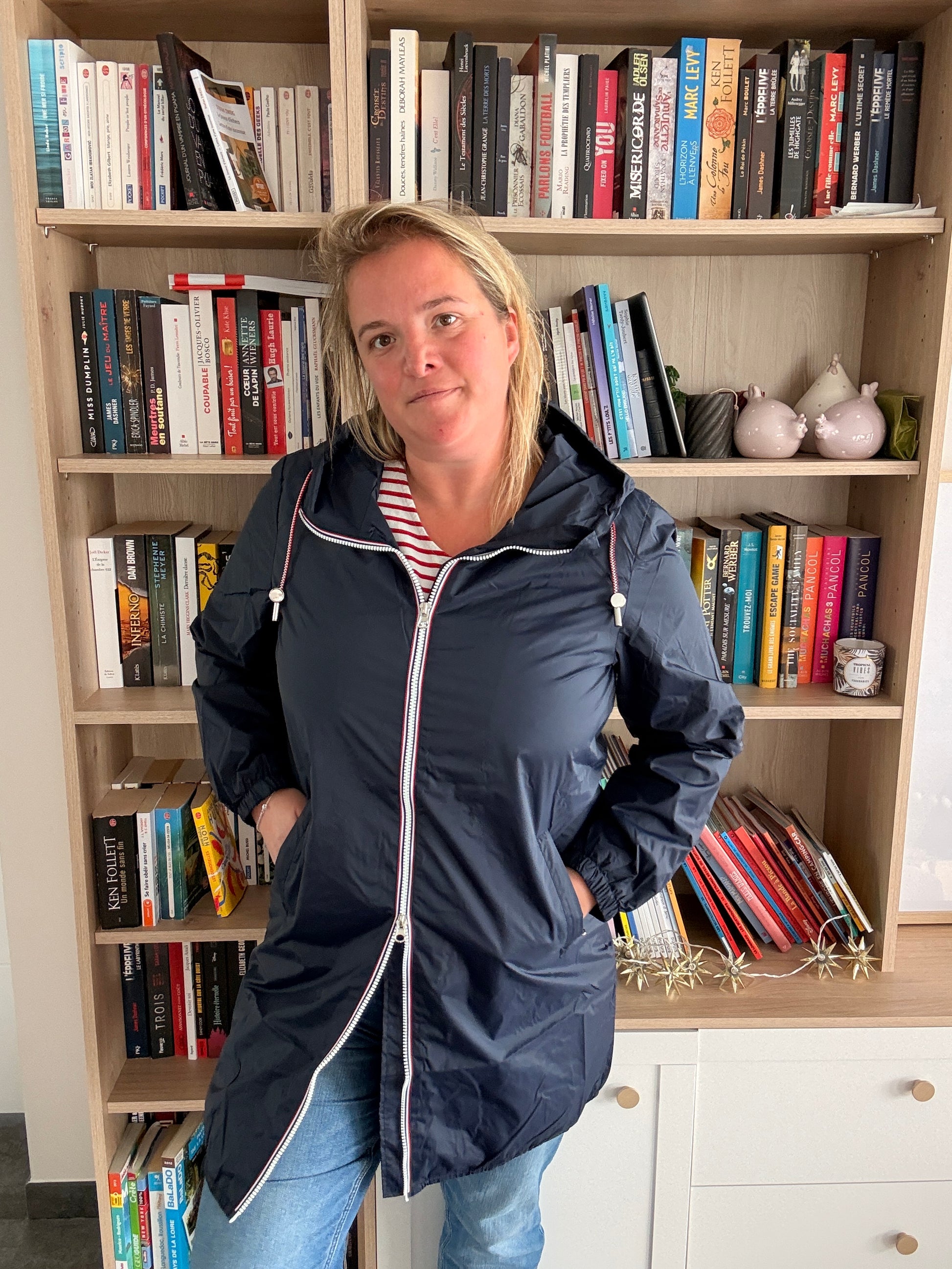 Parka bleu marine imperméable longue, doublure légère, femme debout devant une bibliothèque.
