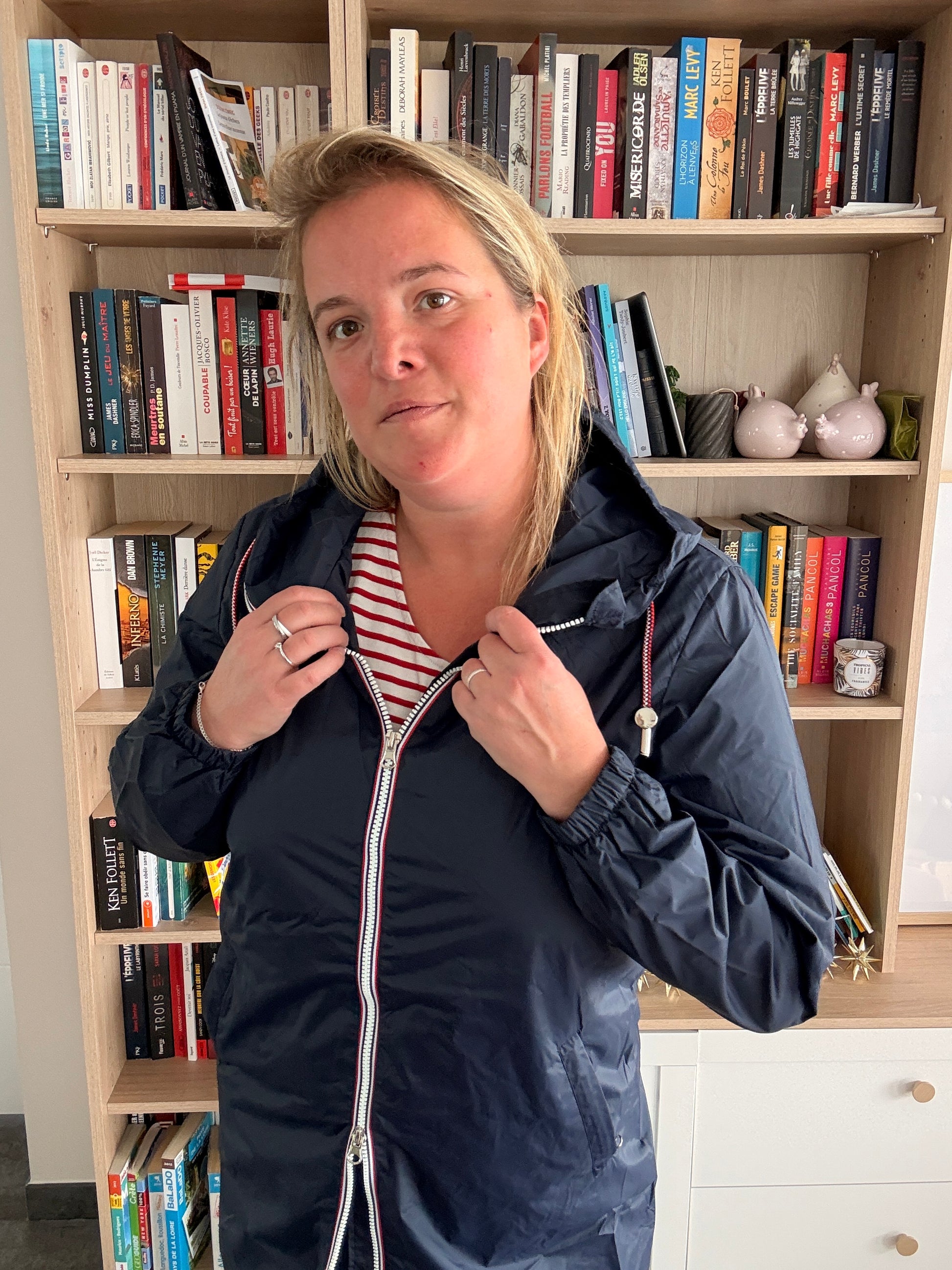 Parka manteau bleu marine imperméable longueur moyenne, avec doublure fine, femme regardant l’appareil, près d'une bibliothèque. Laura Jo.