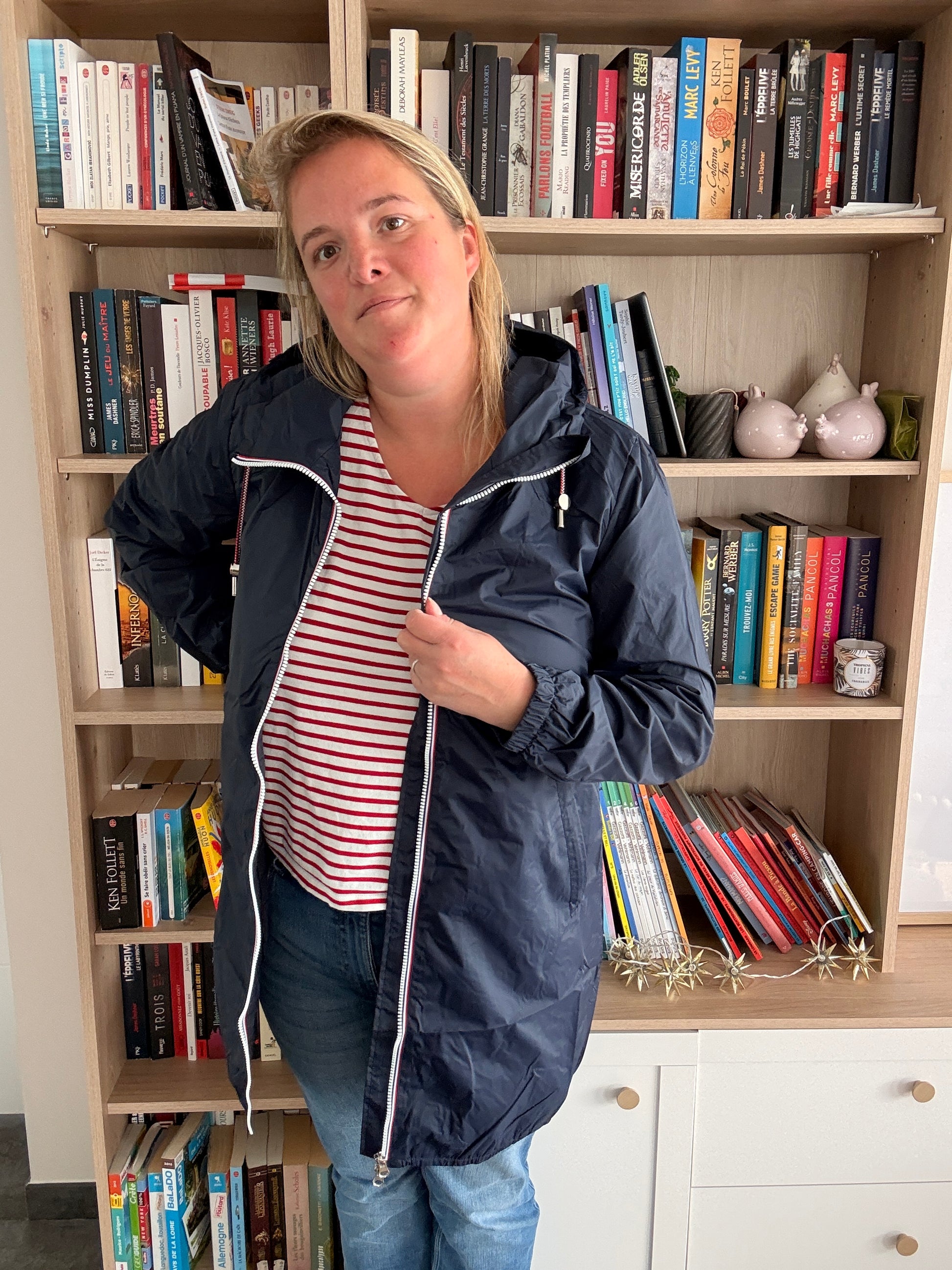 Parka manteau bleu marine imperméable longue avec doublure légère, portée par une femme près d’une étagère remplie de livres.