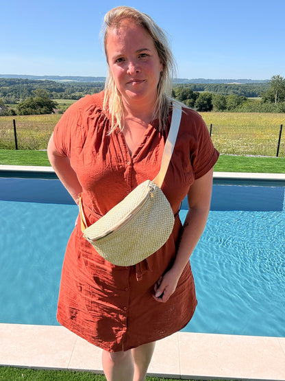 Sac banane en paille porté en mode Bohème sur une robe d’été orange, près d’une piscine extérieure.