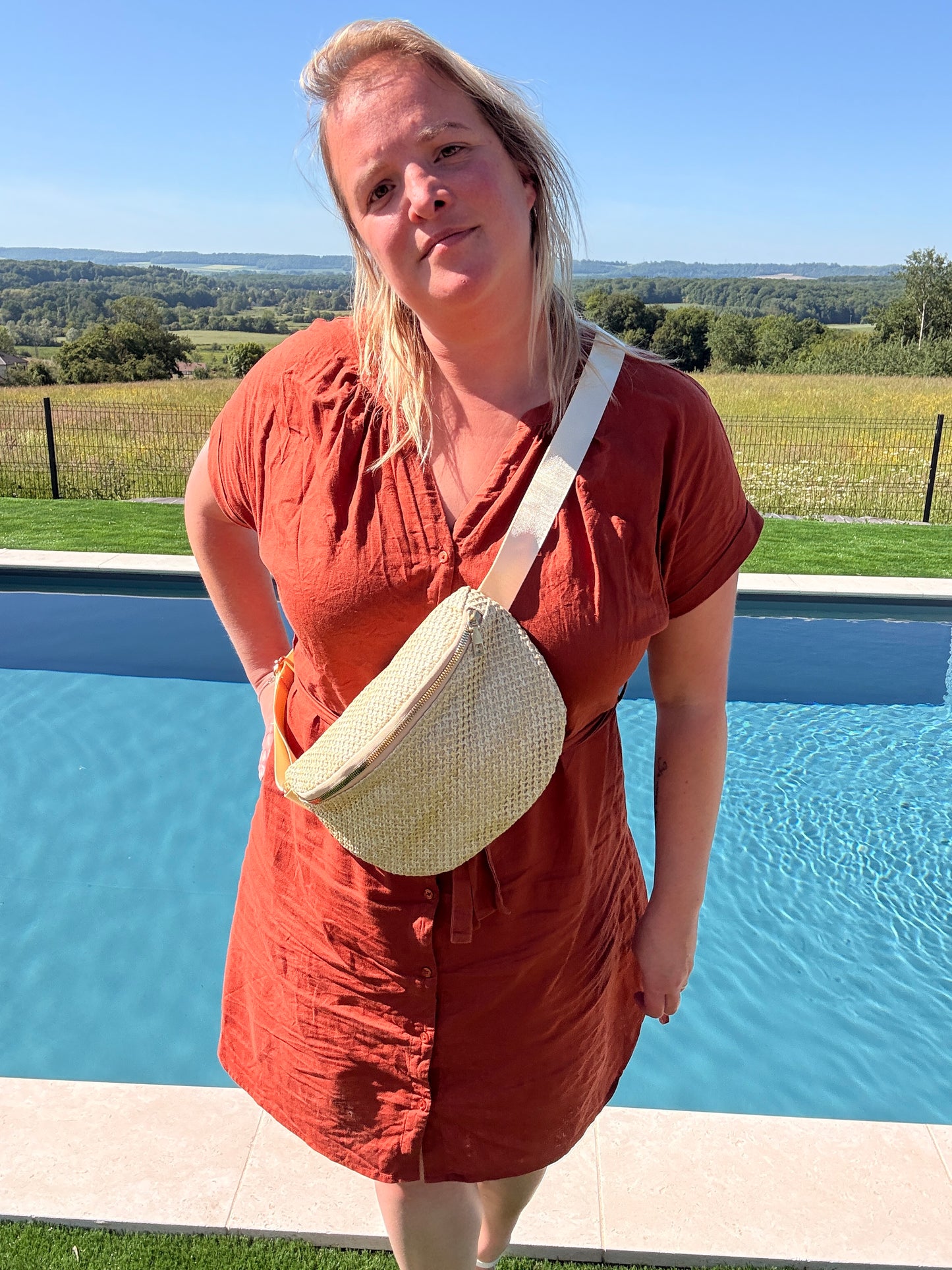Femme souriante porte un sac banane paille autour du torse, vêtue d’une robe rouge-orange près de la piscine en extérieur.
