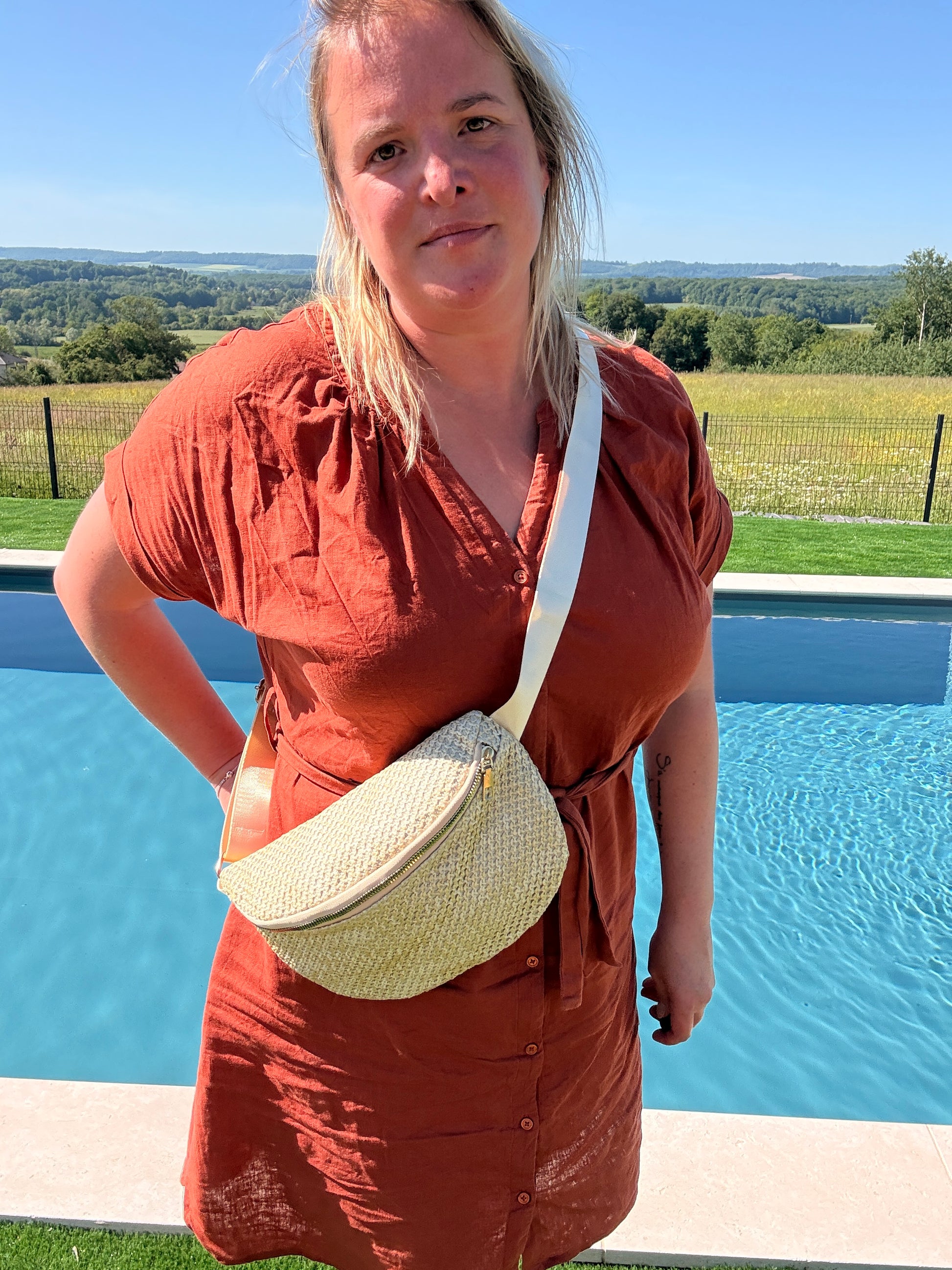Sac banane paille femme porté en style bohème Été: femme près de la piscine, robe orange et sac banane en paille avec bandoulière blanche.