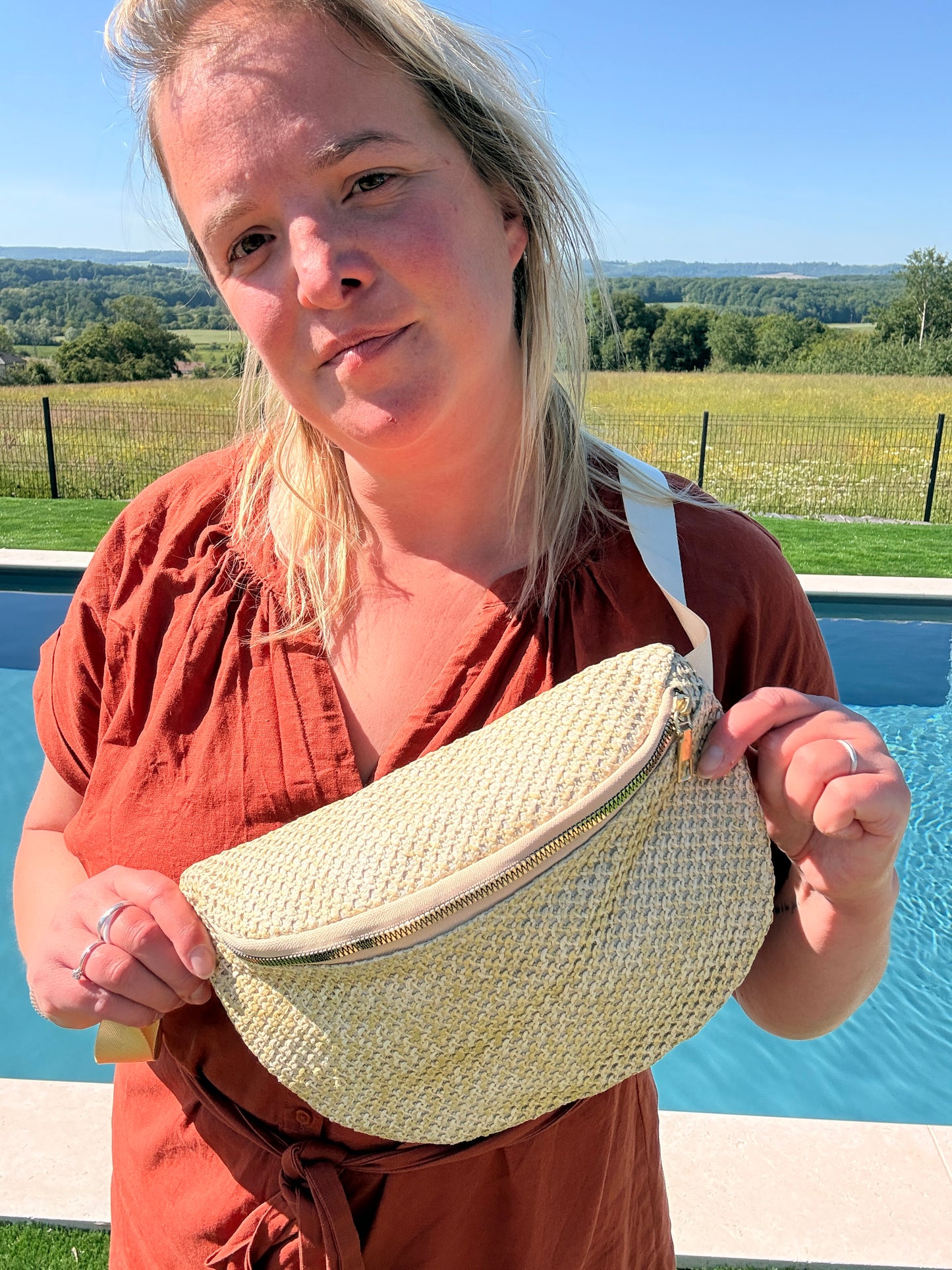 Sac banane paille femme – style bohème été : femme portant une banane tressée beige près de la piscine, paysage champêtre en arrière-plan.