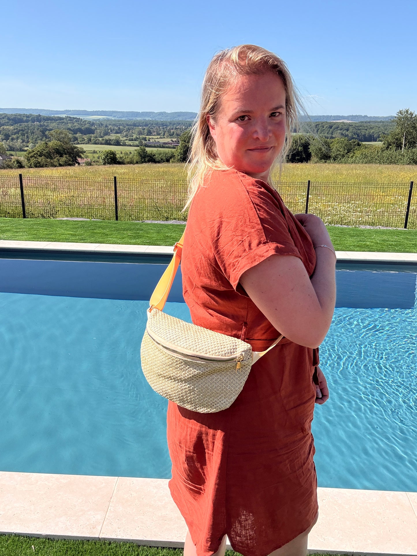 Sac Banane Paille Femme, Style Bohème Été porté en bandoulière par une femme près d’une piscine extérieure, paysage rural en arrière-plan.