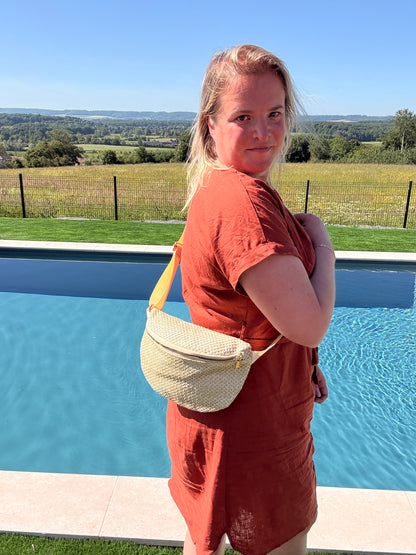 Sac Banane Paille Femme, Style Bohème Été porté en bandoulière par une femme près d’une piscine extérieure, paysage rural en arrière-plan.