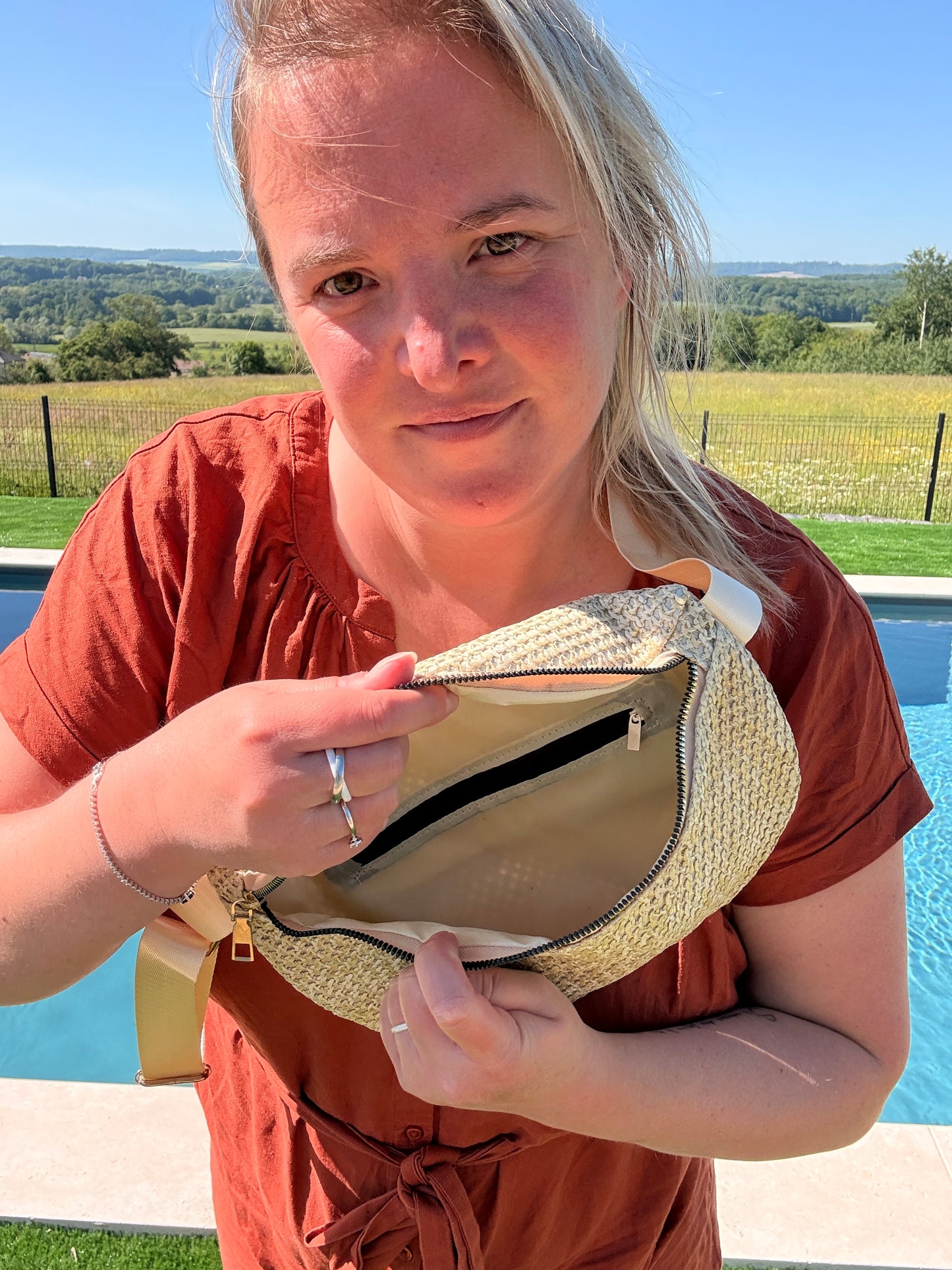 Femme locale près d’une piscine, montrant l’intérieur d’un sac banane en paille beige avec une poche zippée.