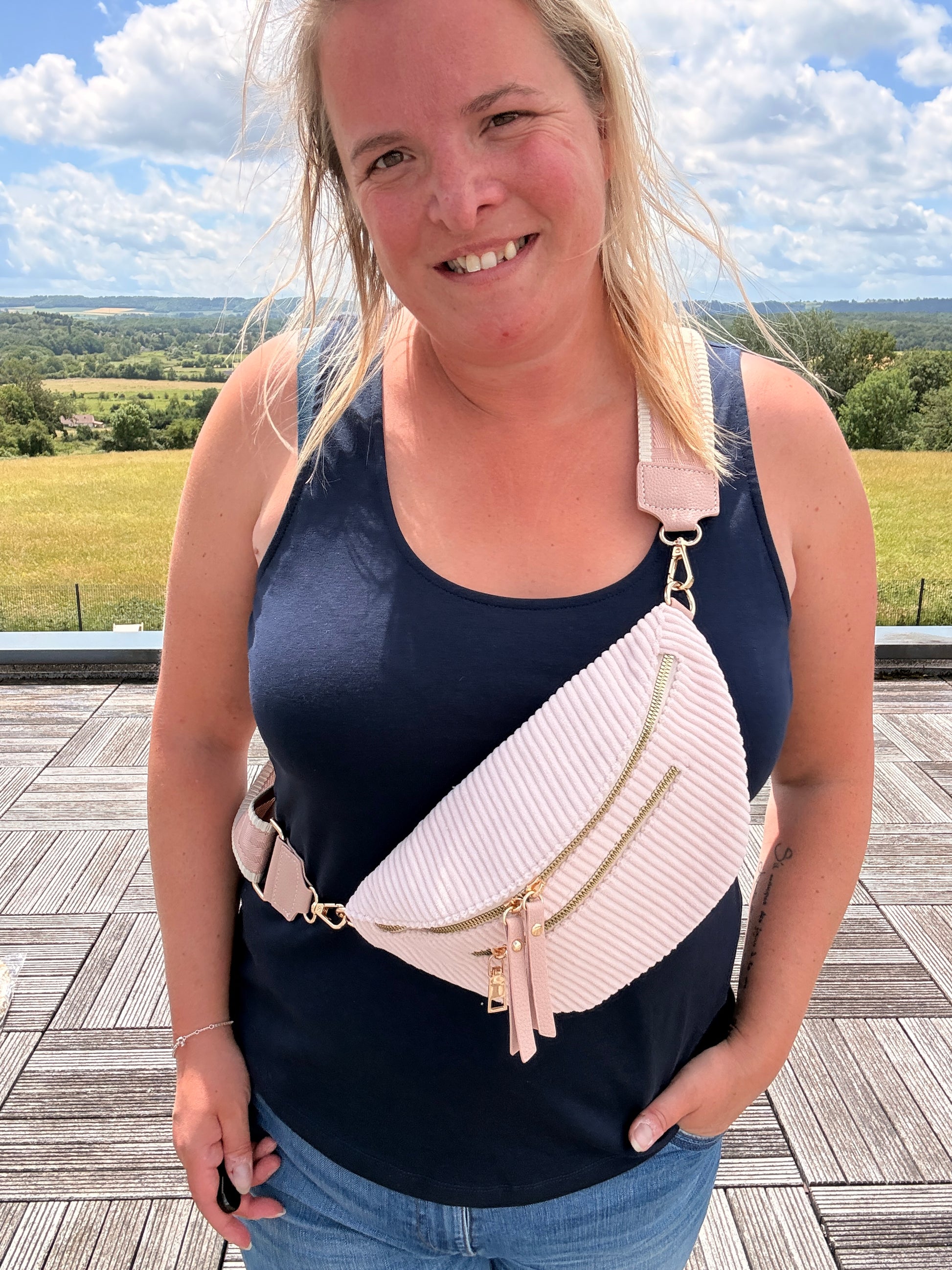 Sac banane velours côtelé femme rose pâle porté en bandoulière; femme souriante sur une terrasse en plein air, avec sangle réglable et deux poches zippées.