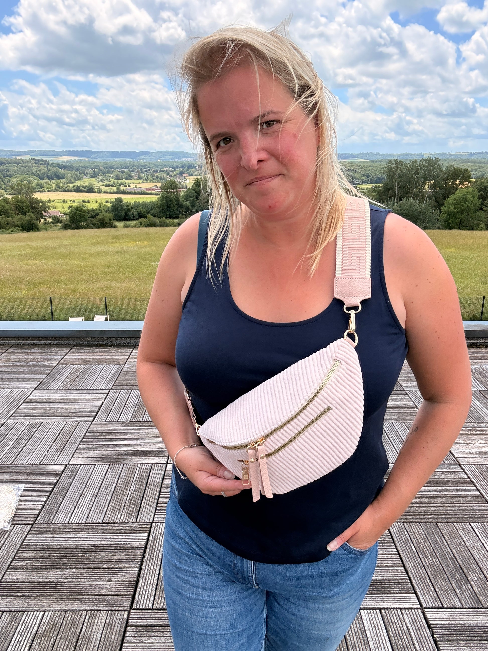 Sac banane velours côtelé rose pâle porté en bandoulière par une femme en débardeur bleu, avec deux poches zippées et sangle réglable.